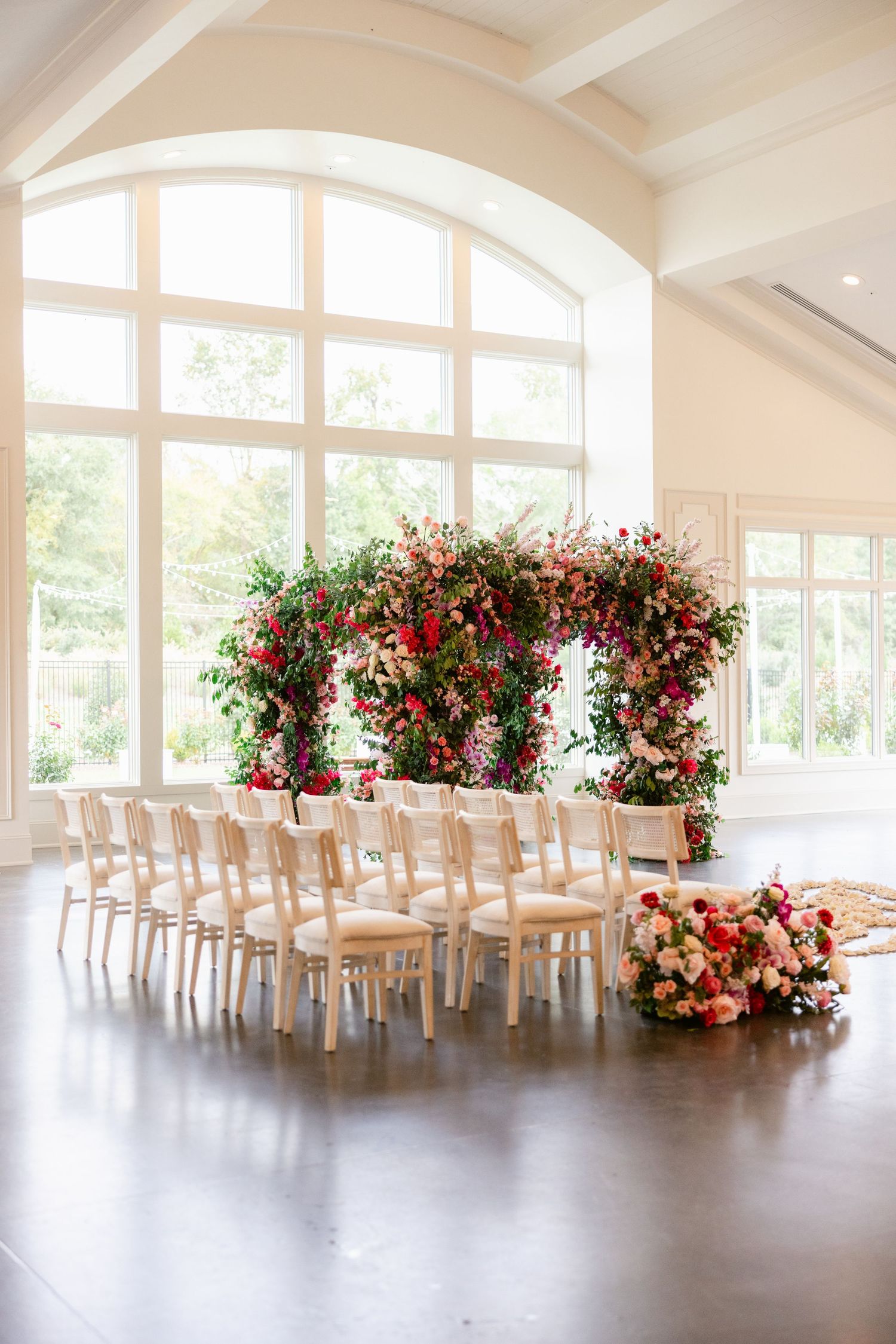 Bright wedding venue with arched windows and red floral arrangements decorating white chairs.