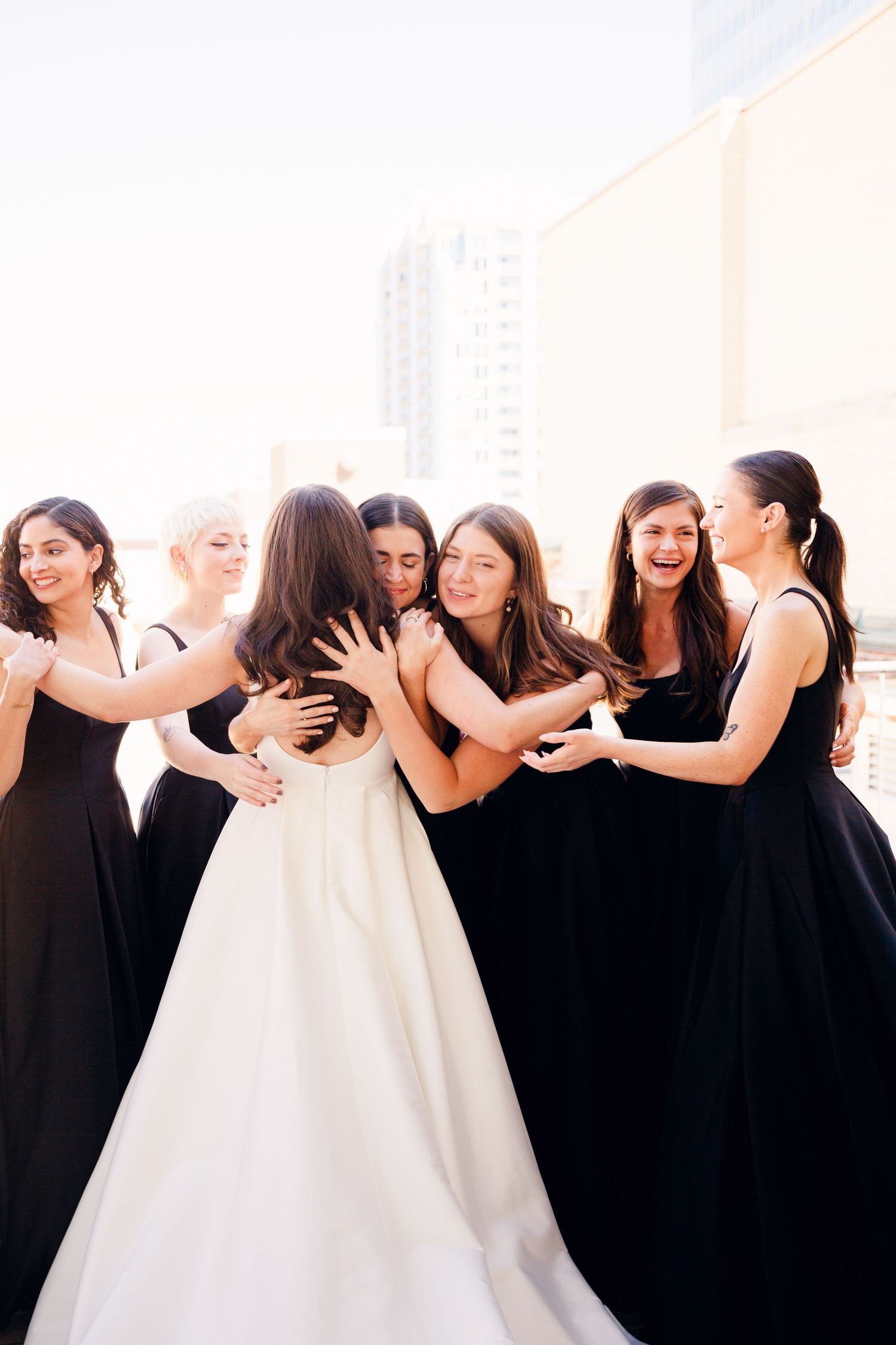A group wearing black dresses embraces in a tender moment on a rooftop setting.