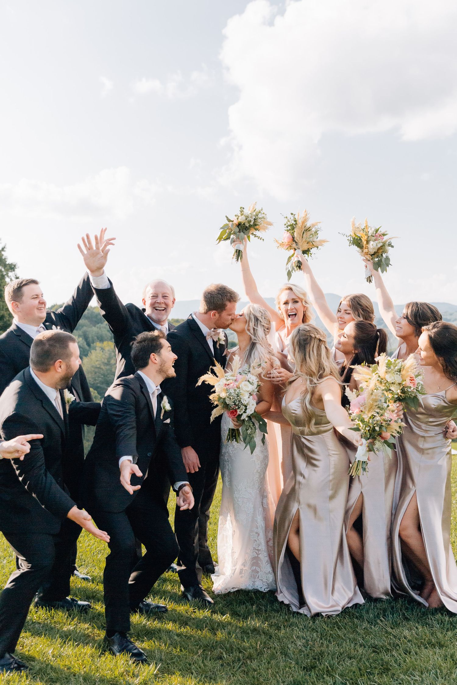 Wedding party celebrates joyfully on a hilltop with bridesmaids in champagne dresses holding floral bouquets.