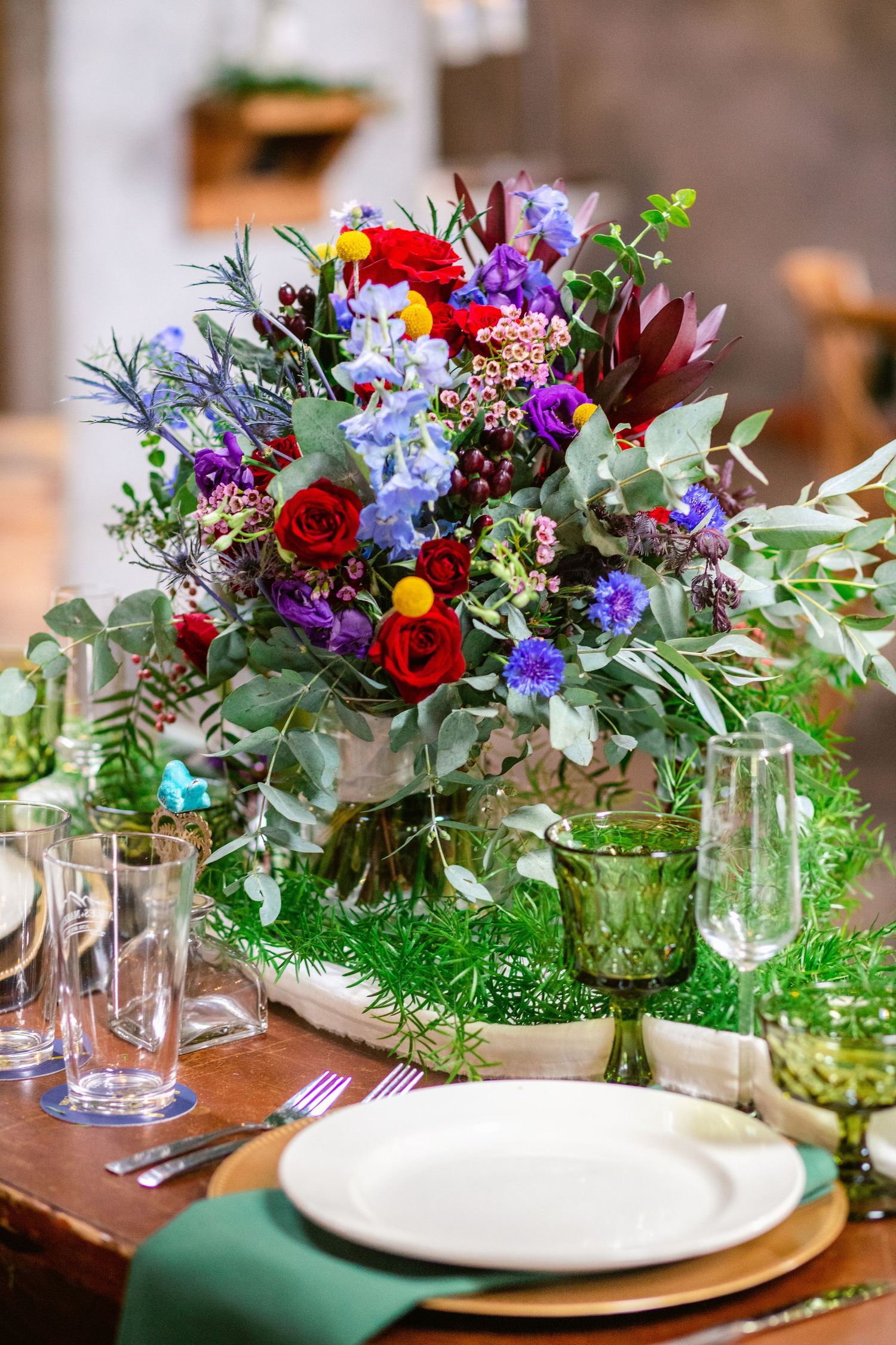 Vibrant wedding centerpiece with red roses, purple flowers, and greenery on a rustic table setting.