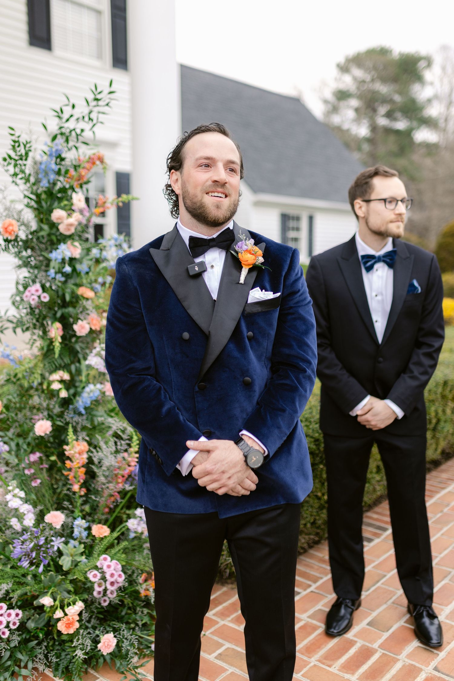 Wedding party members in formal attire stand by floral arrangement with navy velvet tuxedo and black bow ties.