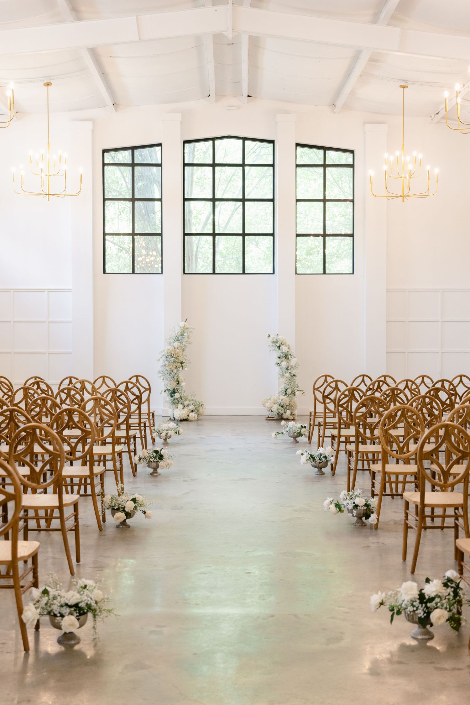 White wedding ceremony space with arched windows, golden chairs lined up in rows, and delicate floral arrangements along the aisle.