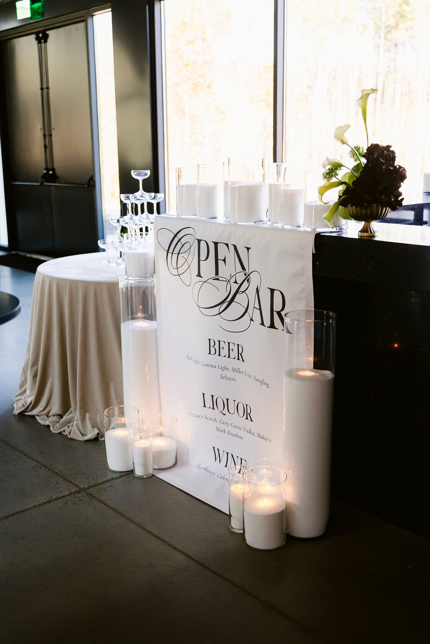 Bar setup with white signage for beer, wine and liquor displayed alongside white candles on dark surface.