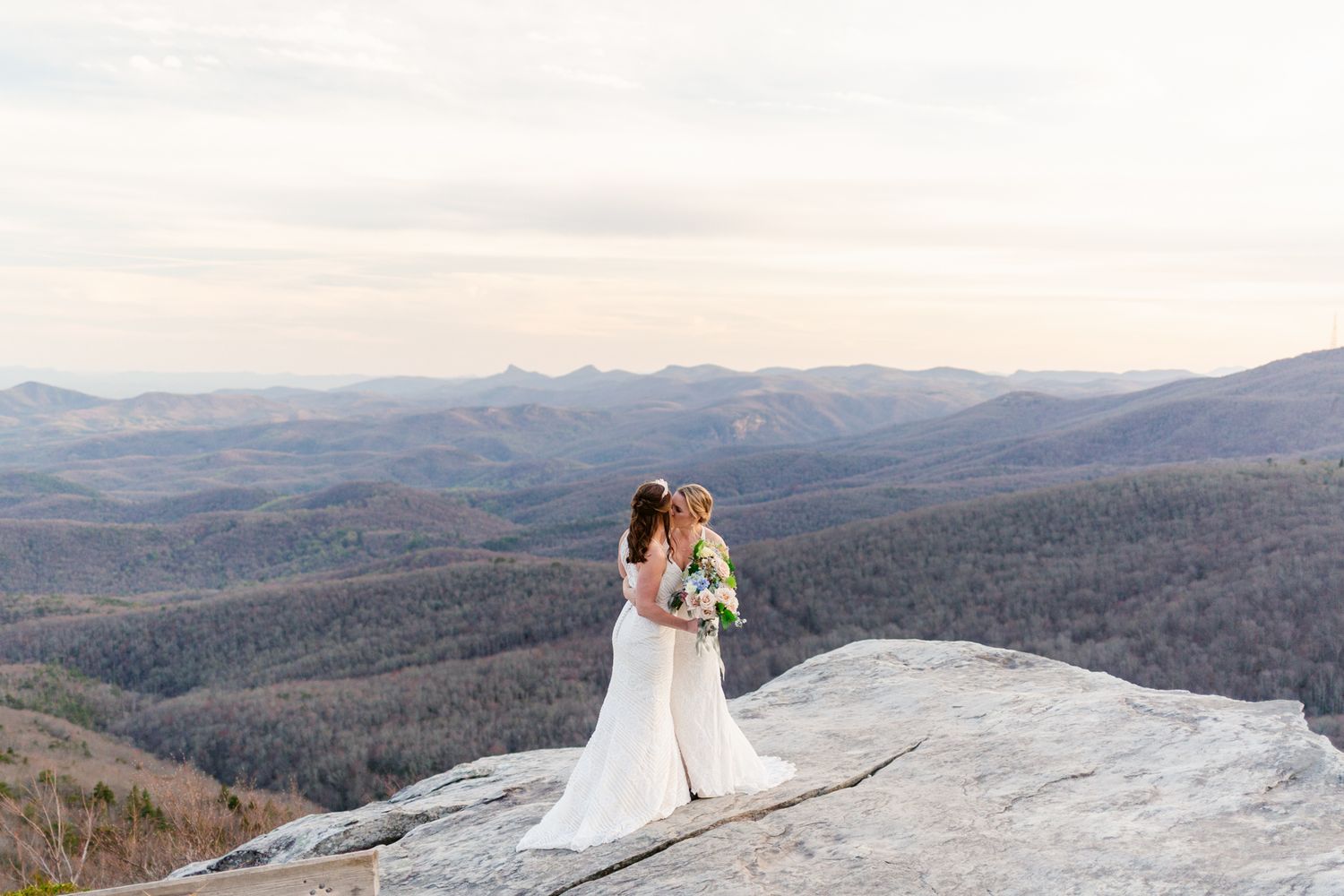 A couple in wedding attire stands atop a scenic mountain overlook with rolling hills stretching into the distance.