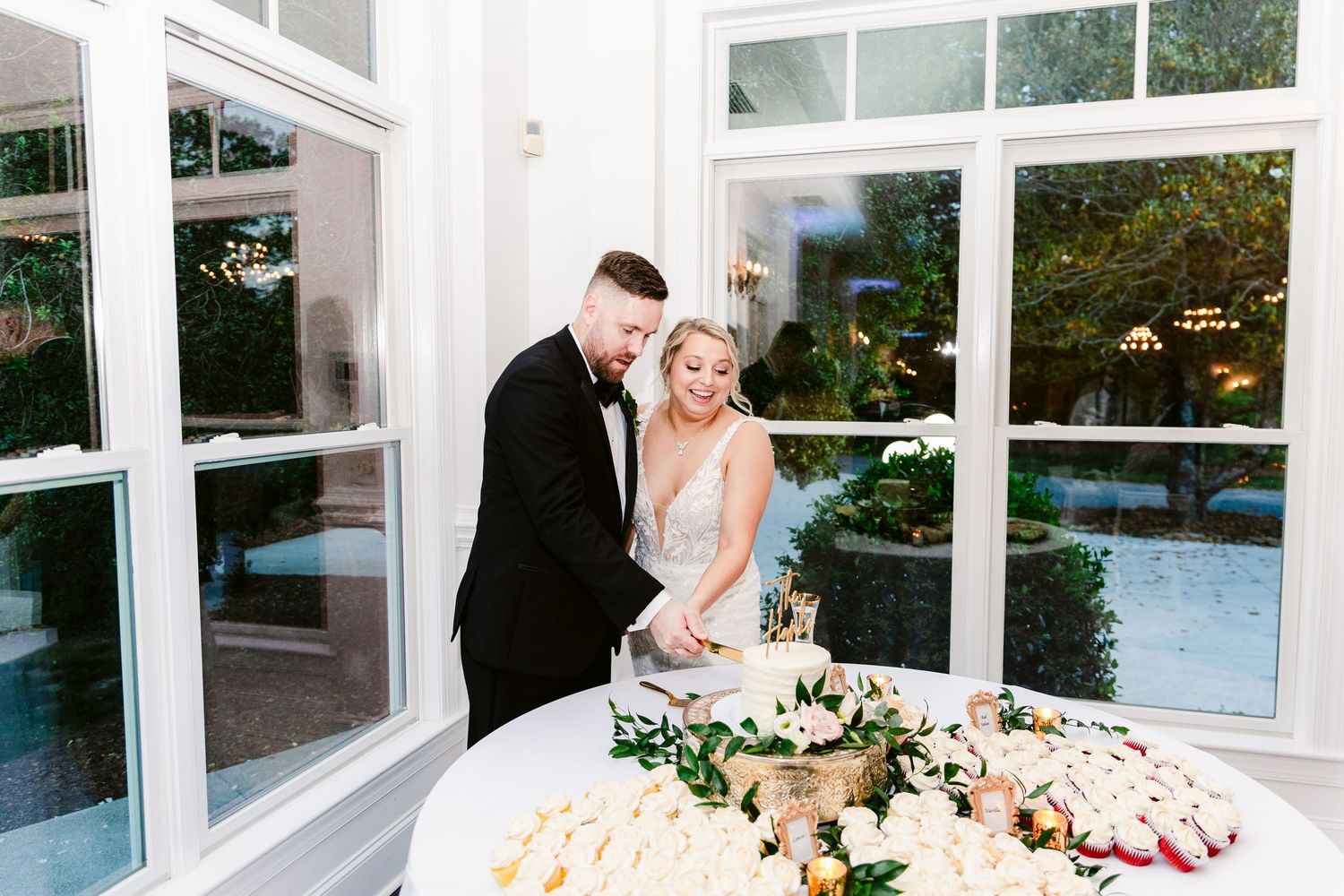 A wedding reception venue features elegant white decor and floral arrangements on dining tables near large windows.