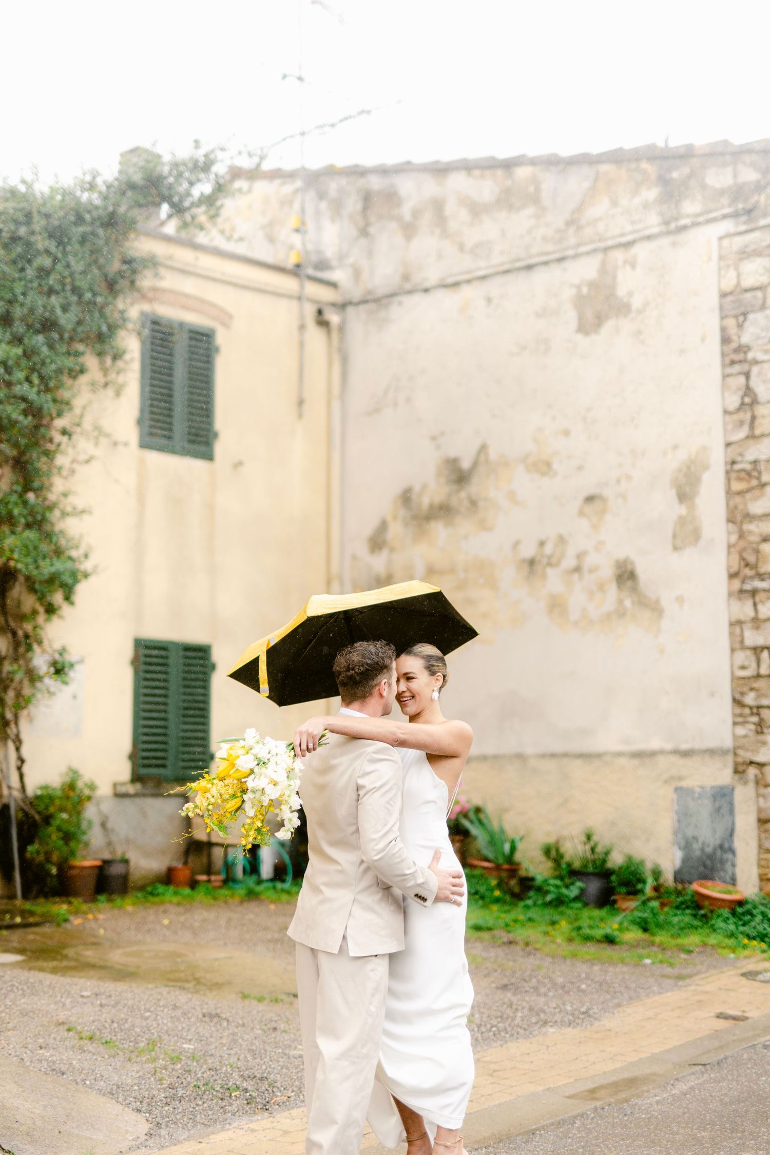 A couple in white outfits share a joyful embrace in a rustic courtyard setting with weathered walls and green shutters.