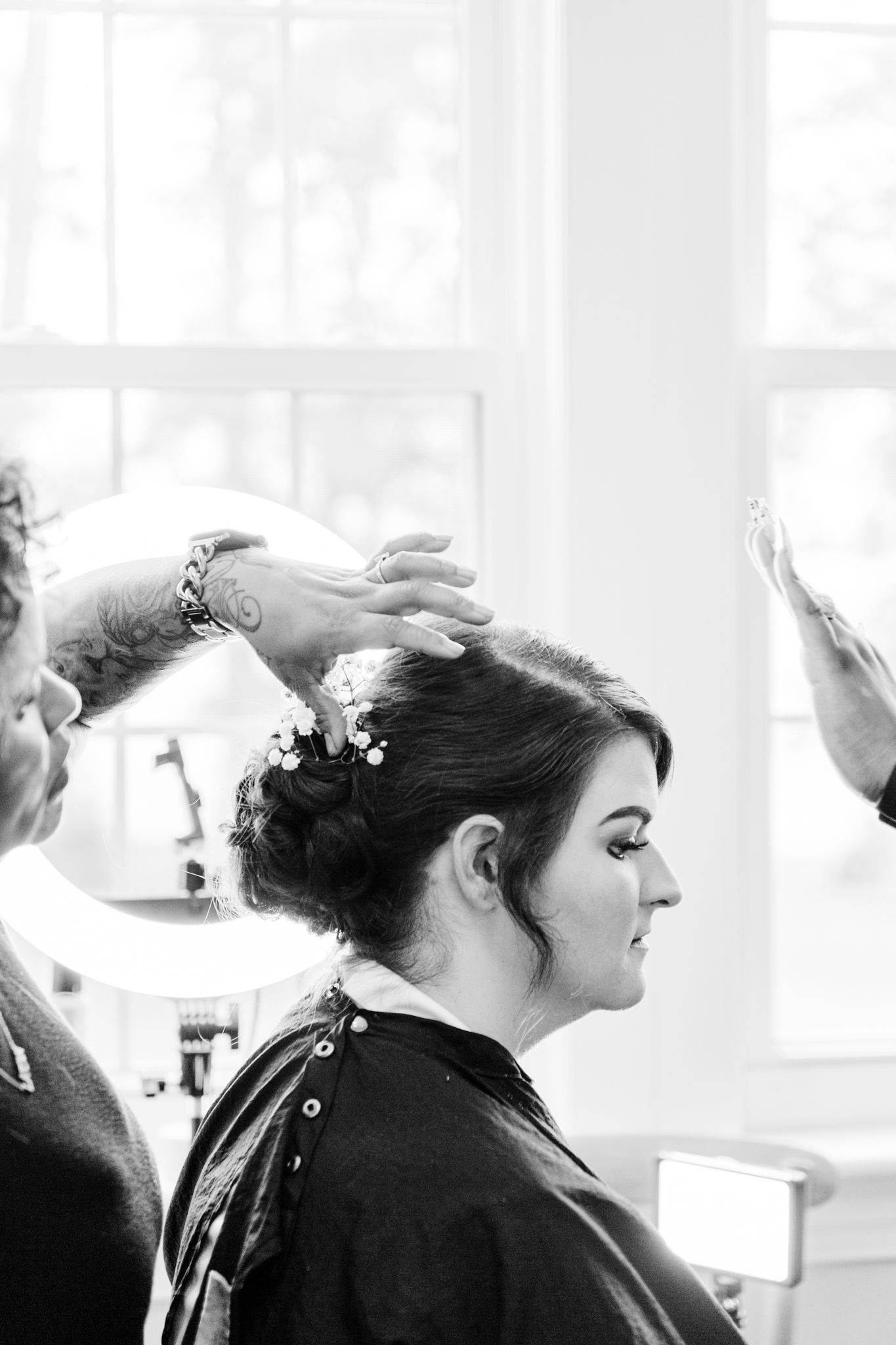 Black and white photo showing hairstylists styling a client's updo in a bright salon setting.