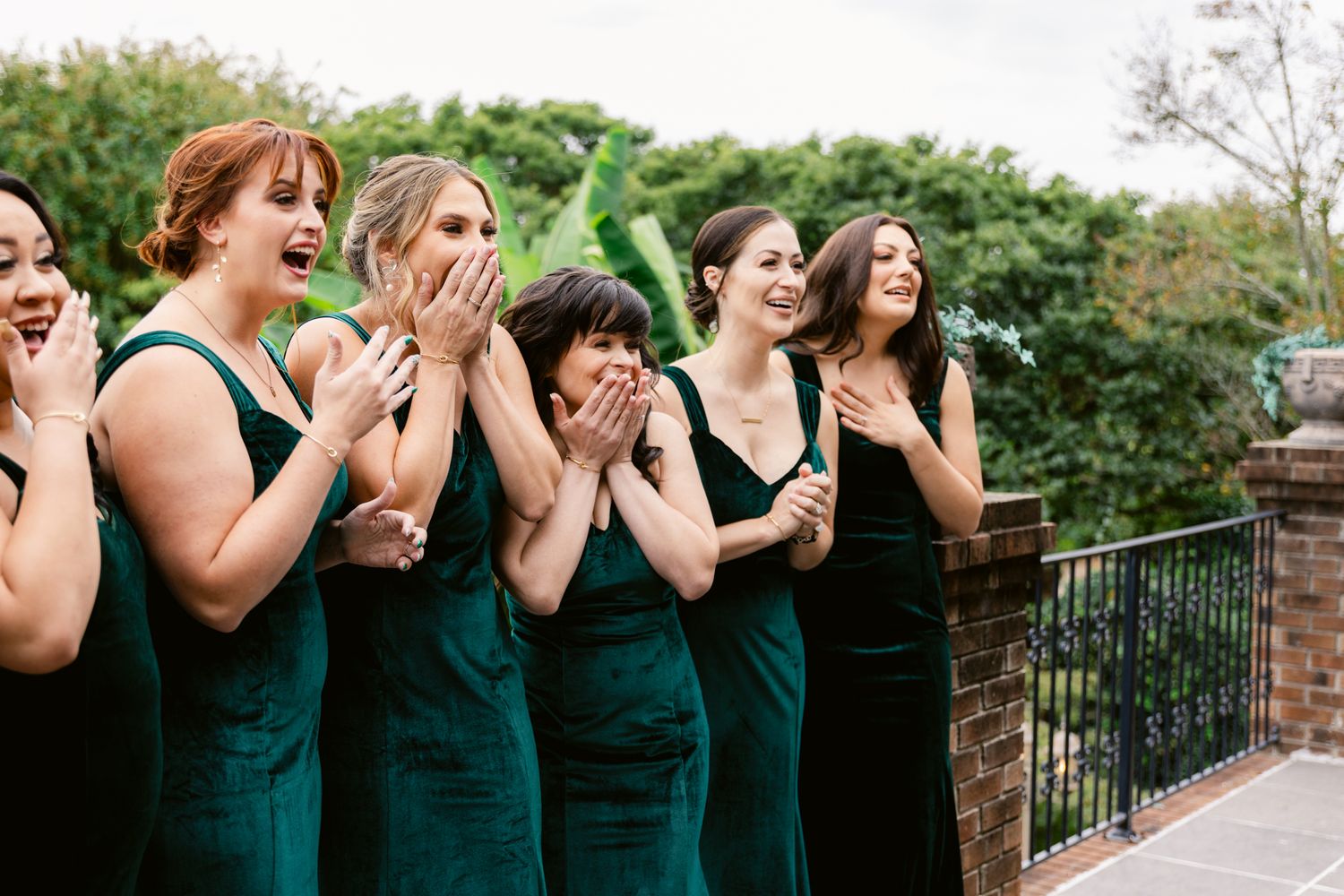 A group of bridesmaids in emerald green dresses react with excitement and joy during a wedding celebration.