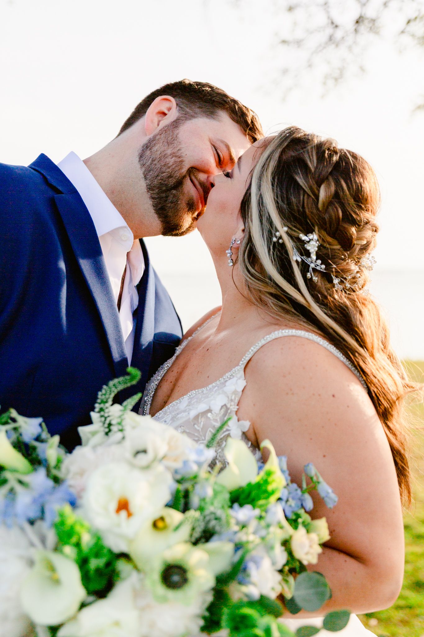 Romantic wedding couple shares intimate kiss at sunset while holding elegant white floral bouquet.