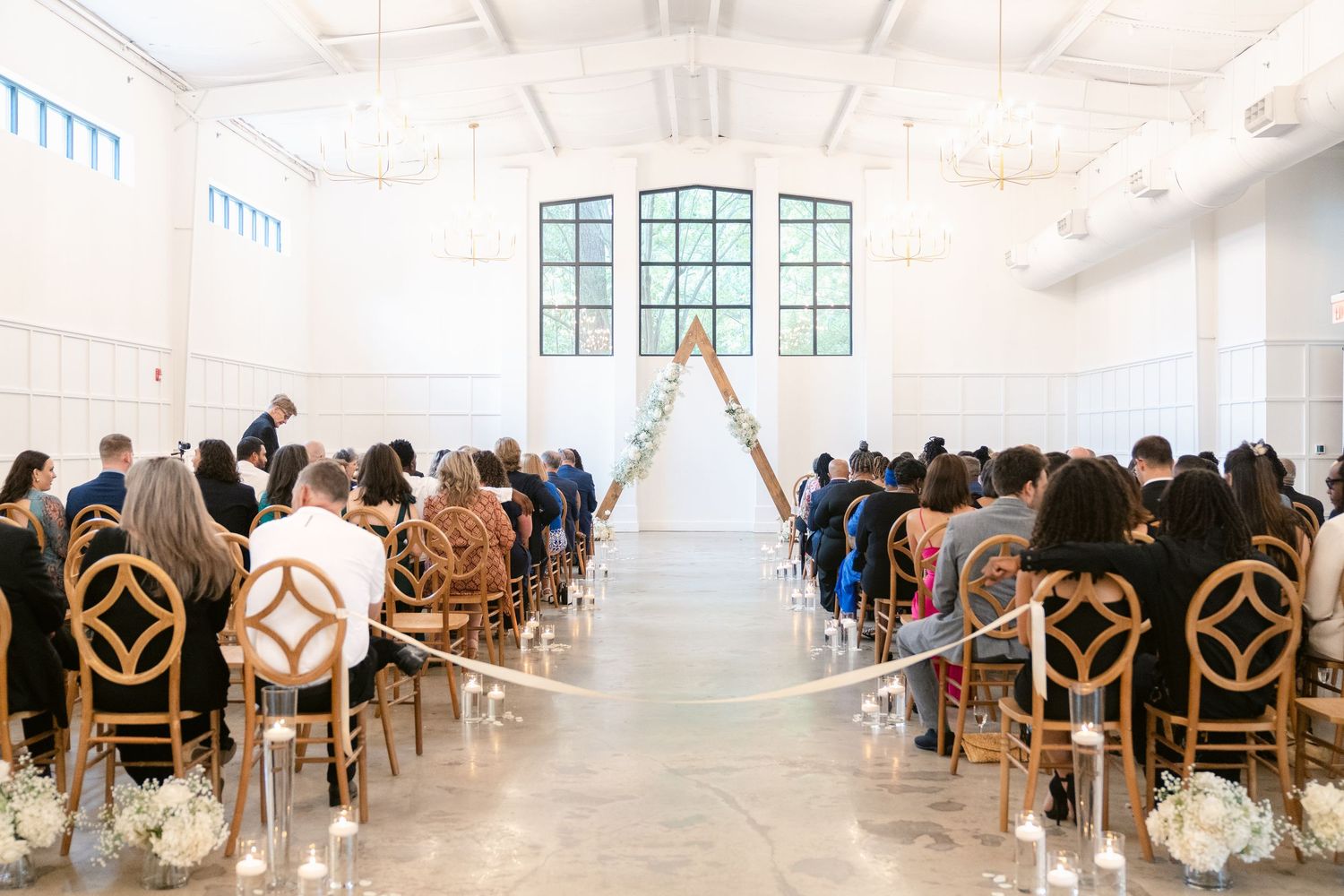 Elegant wedding ceremony setup in a bright white venue with wooden chairs and a center aisle leading to arched windows.