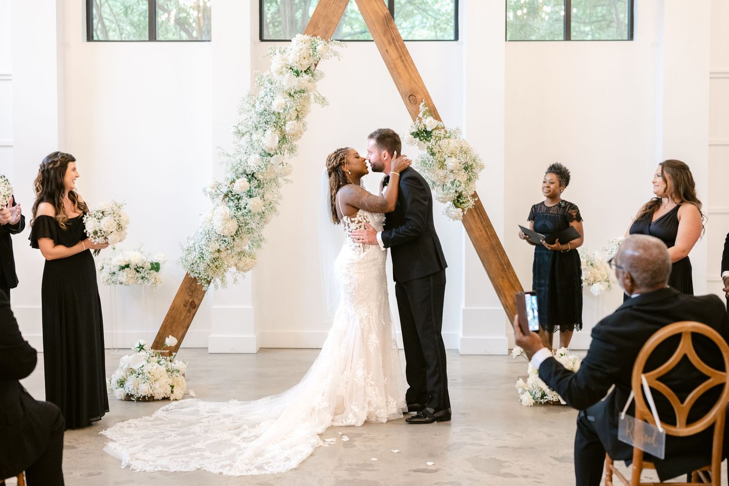 A wedding ceremony unfolds in a bright white room with large windows and floral decorations along the walls.