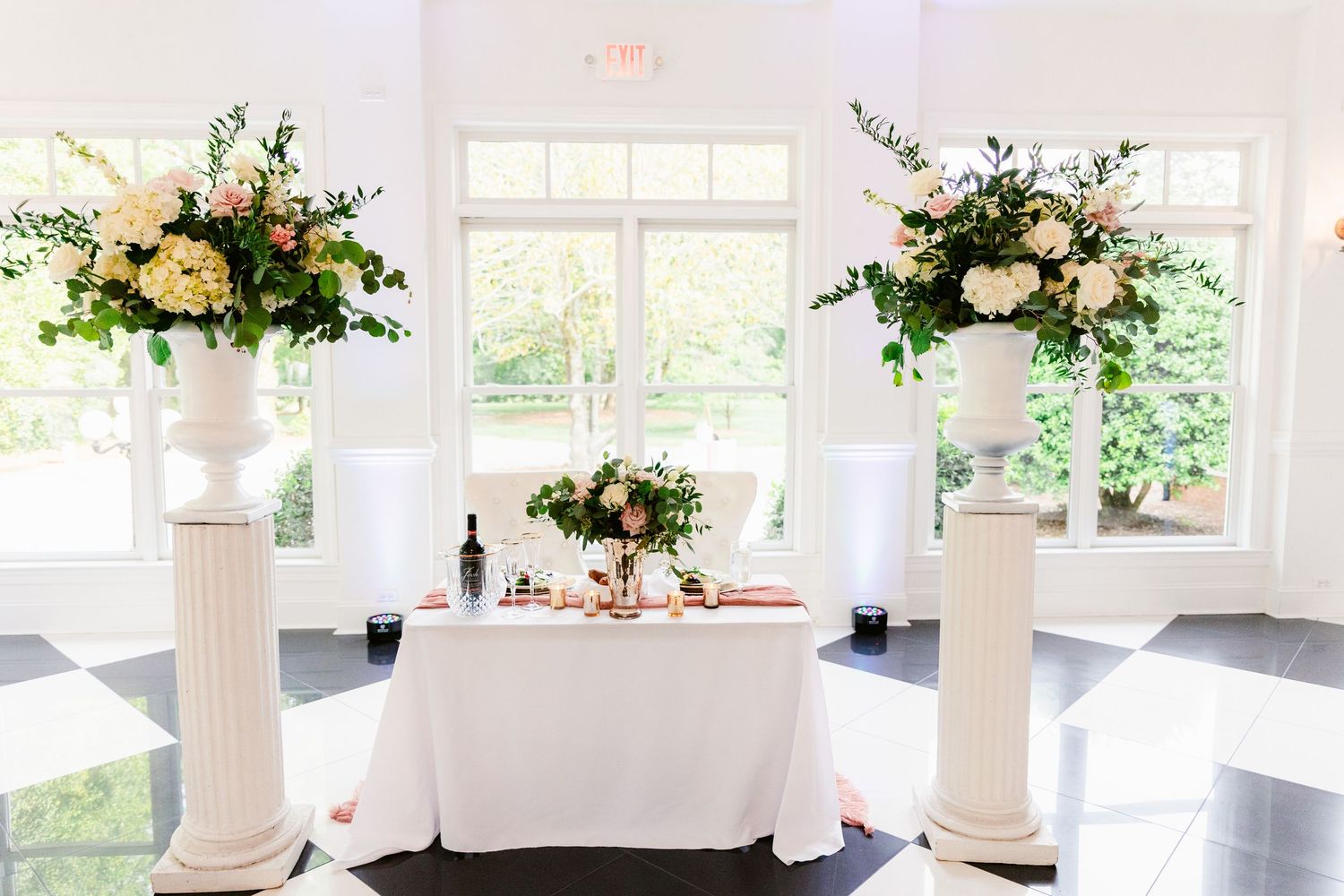 White wedding reception venue with tall columns, draped fabric, and lush floral arrangements on round tables.