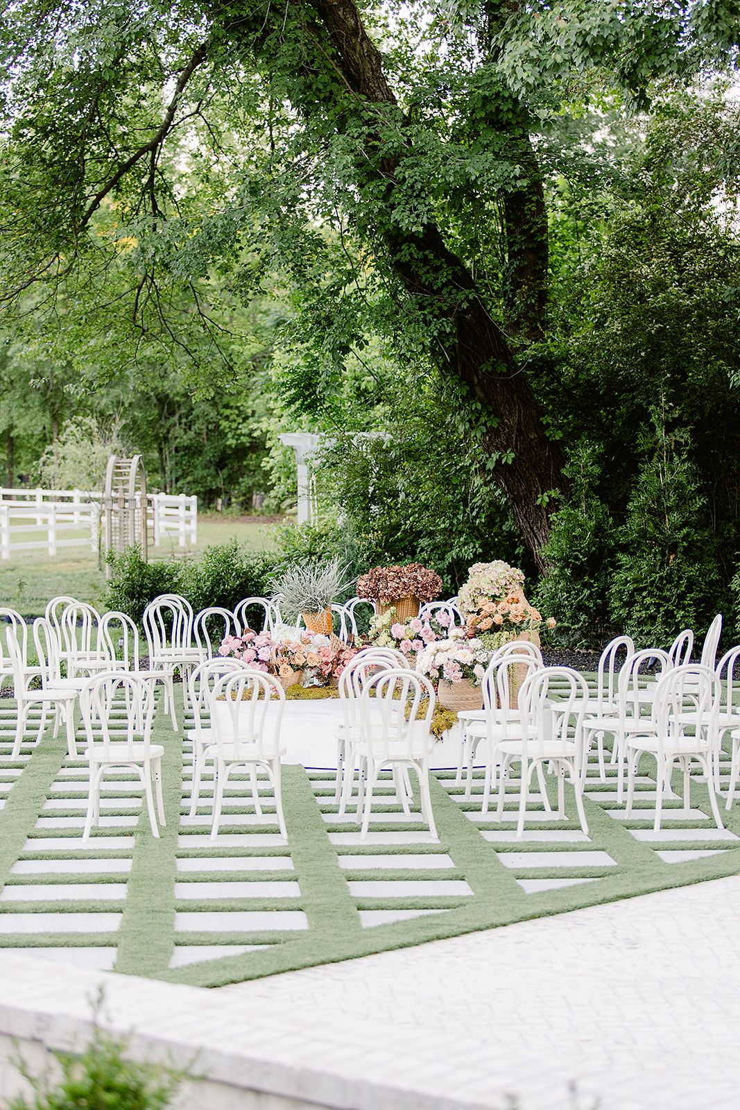 Outdoor wedding ceremony setup with rows of white chairs on grass under shady trees and a white fence backdrop.