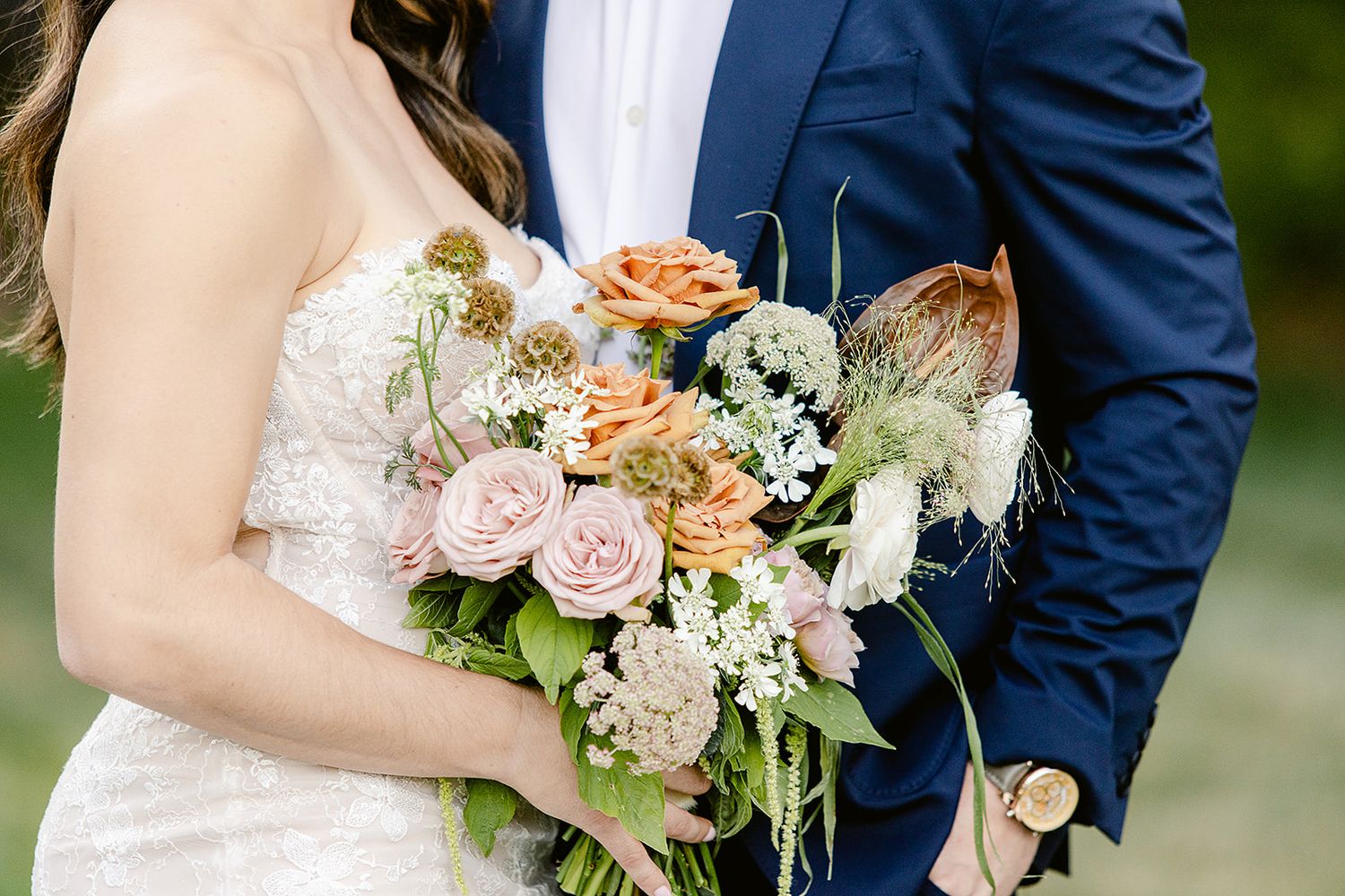 A couple in wedding attire stands close together holding a romantic peach and white floral bouquet outdoors.