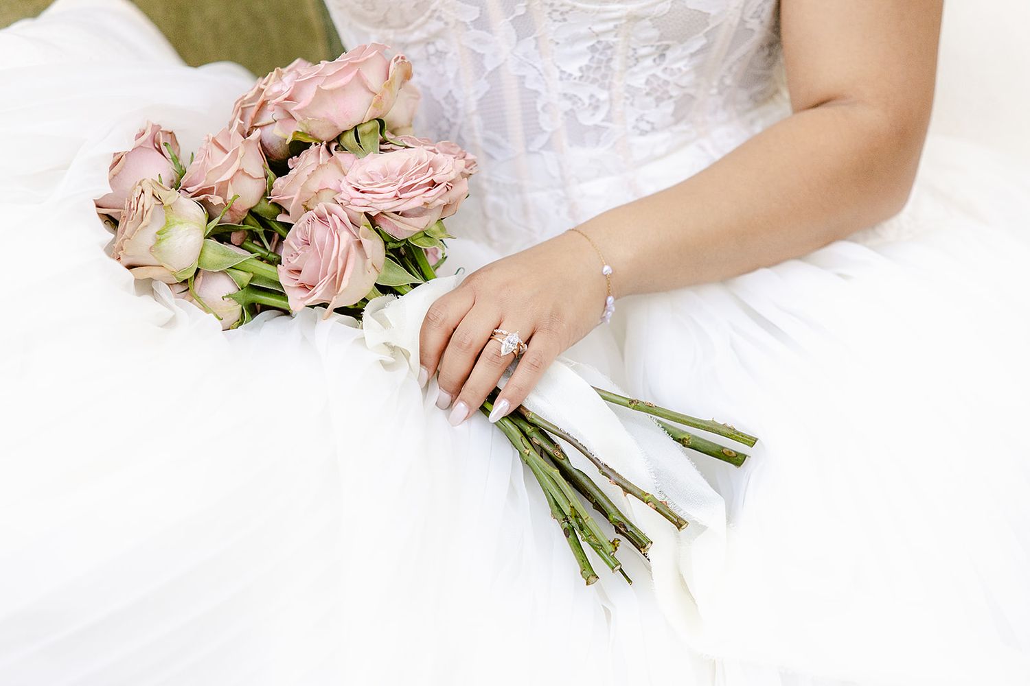 A delicate pink rose bridal bouquet held against a white wedding dress with a manicured hand displaying a ring.