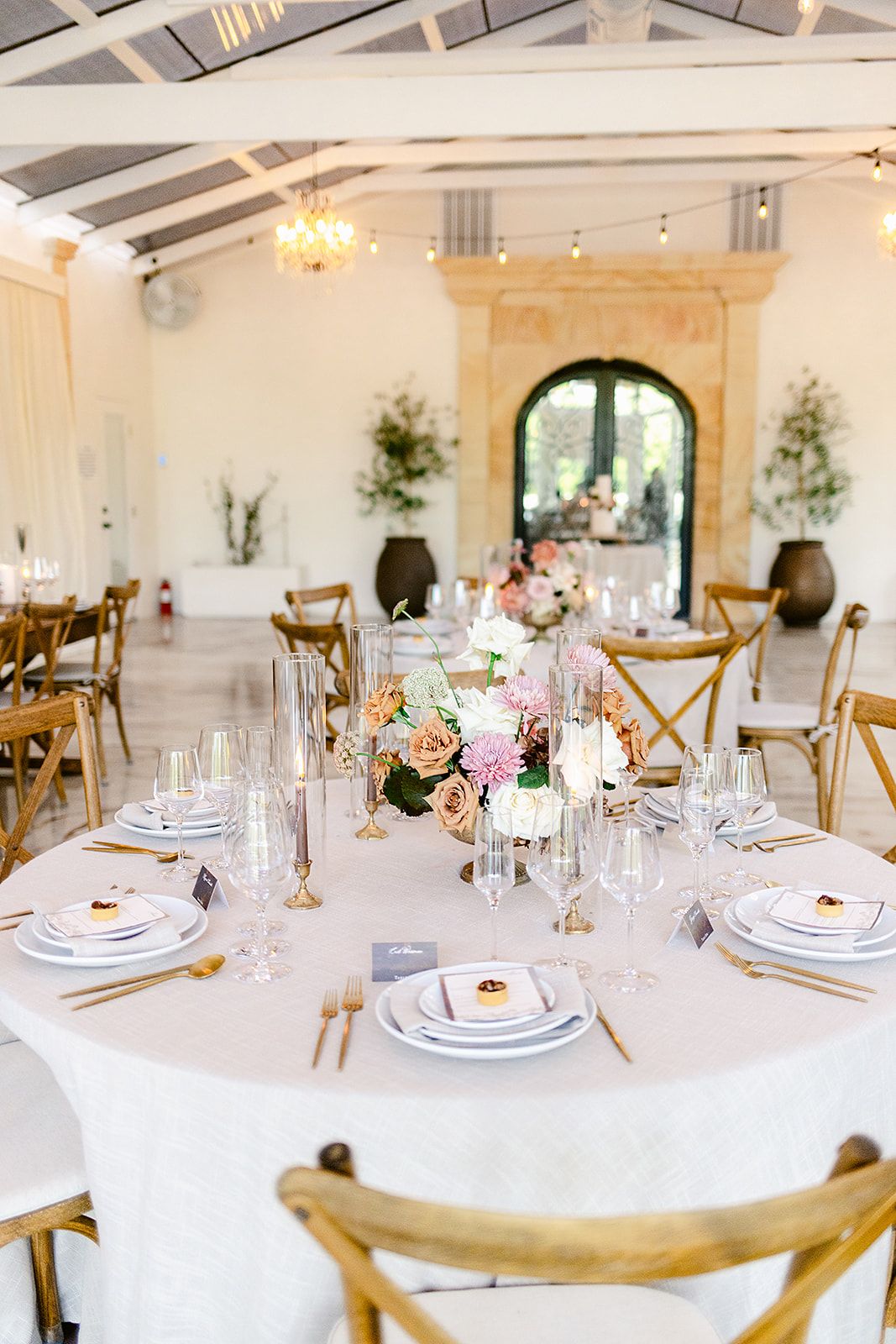 Elegant wedding reception tables set with white linens, crystal glassware, and floral centerpieces in a bright white venue.