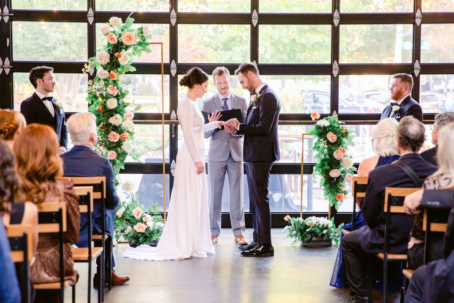 A wedding ceremony takes place in a modern venue with large windows and floral arrangements.