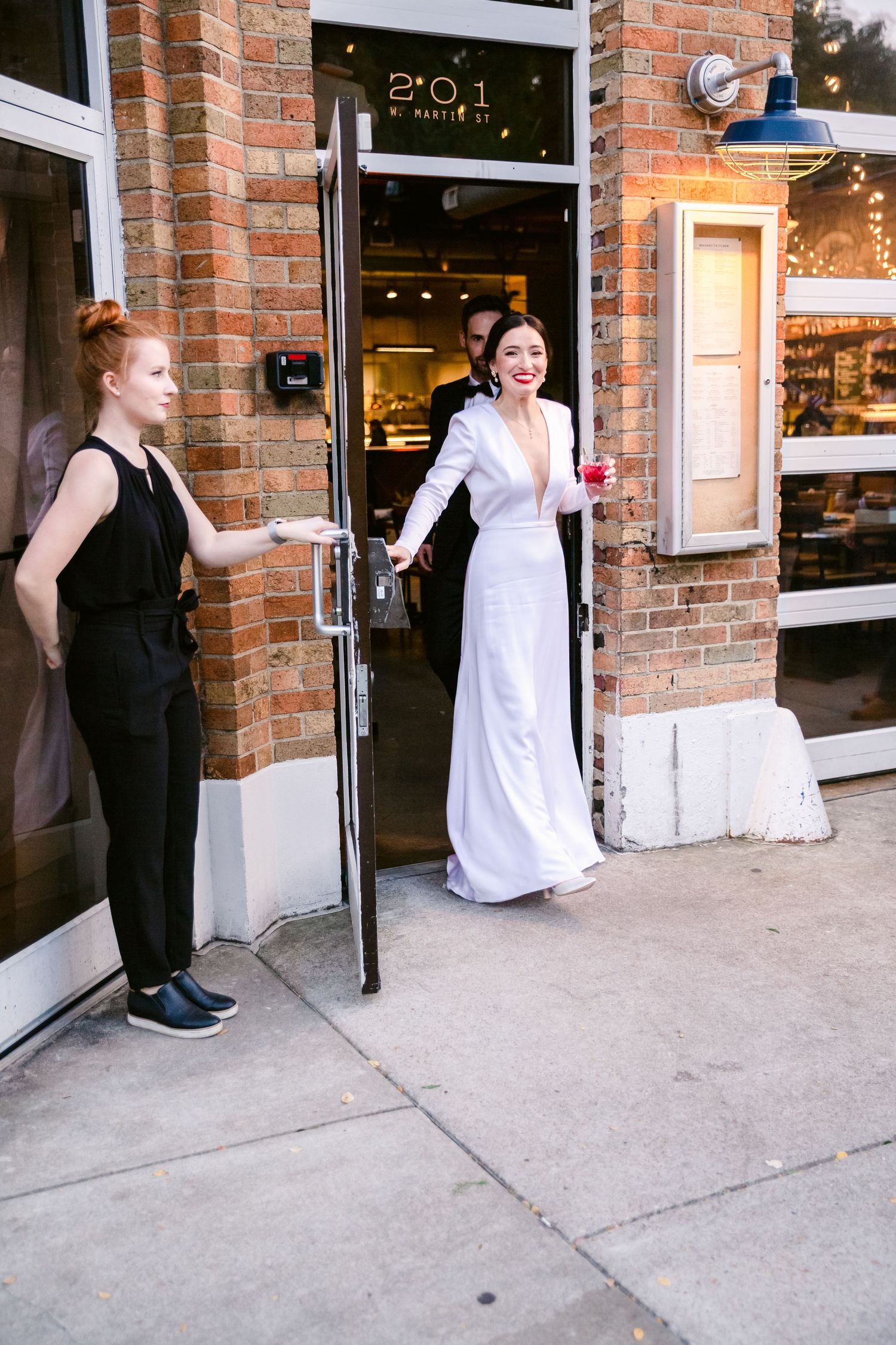 Two people in formal attire playfully interact outside a brick storefront entrance.