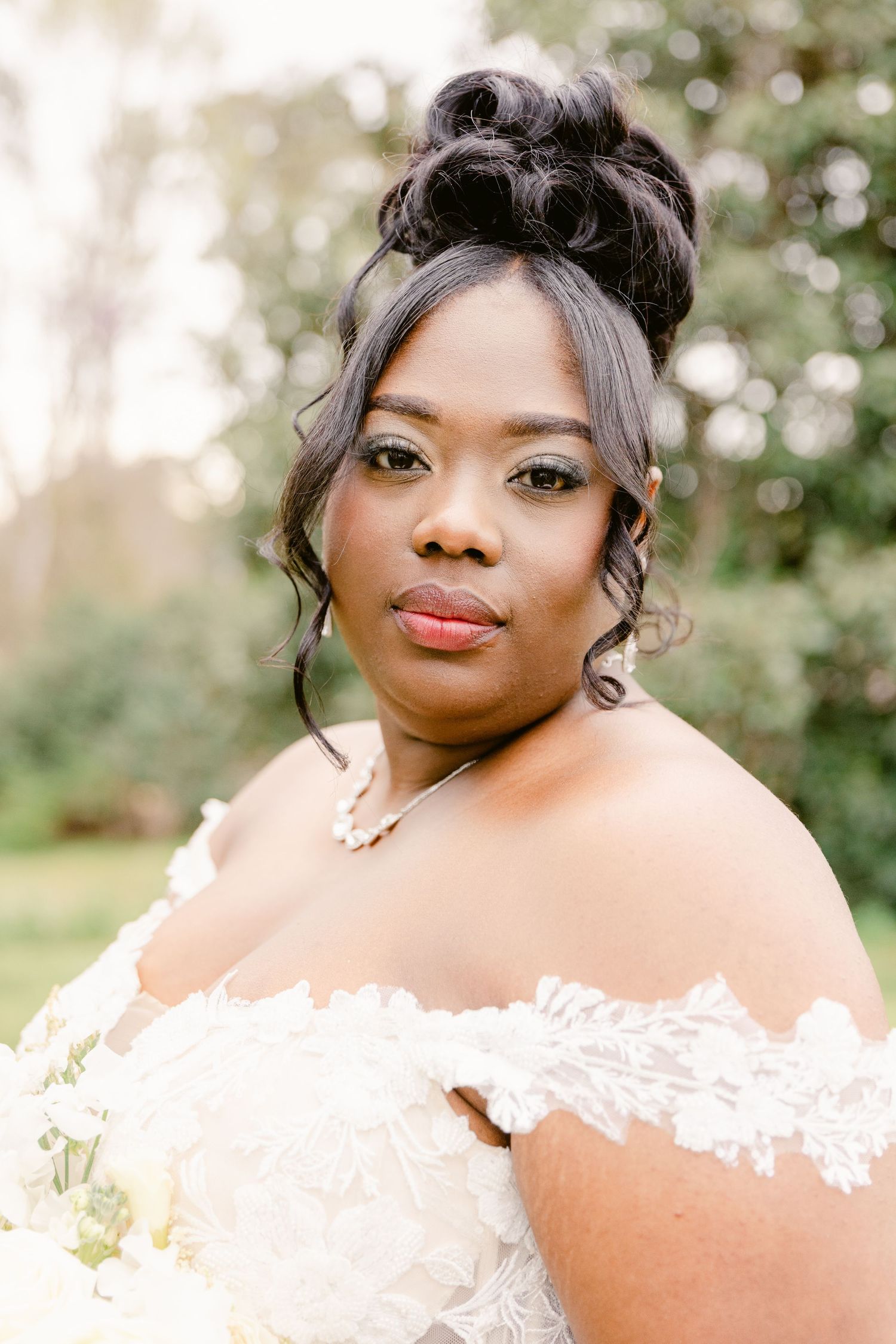 A bride in a white off-shoulder wedding dress poses outdoors with soft natural lighting and foliage in the background.