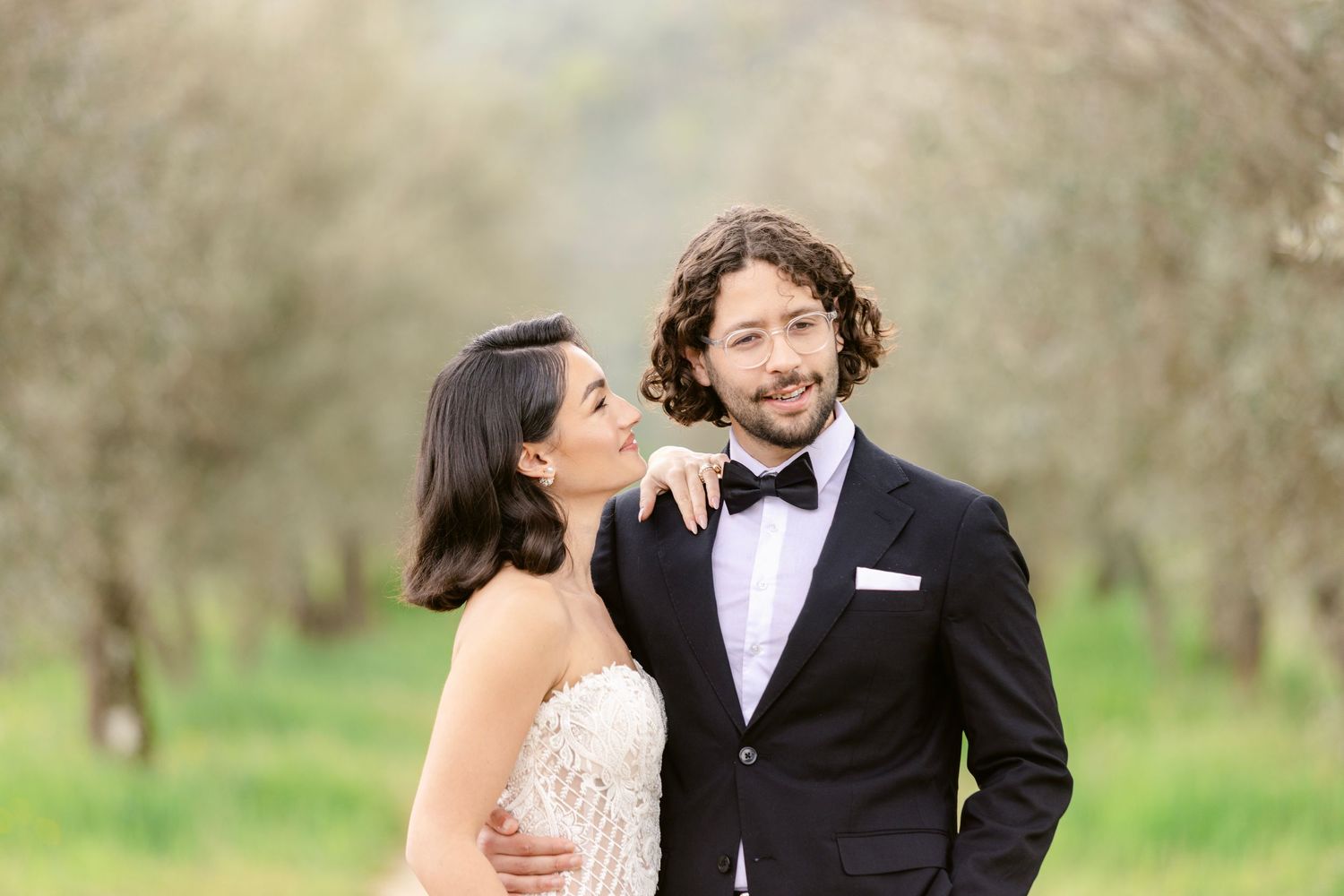 A couple shares a romantic moment in formal attire while standing in a lush spring orchard.