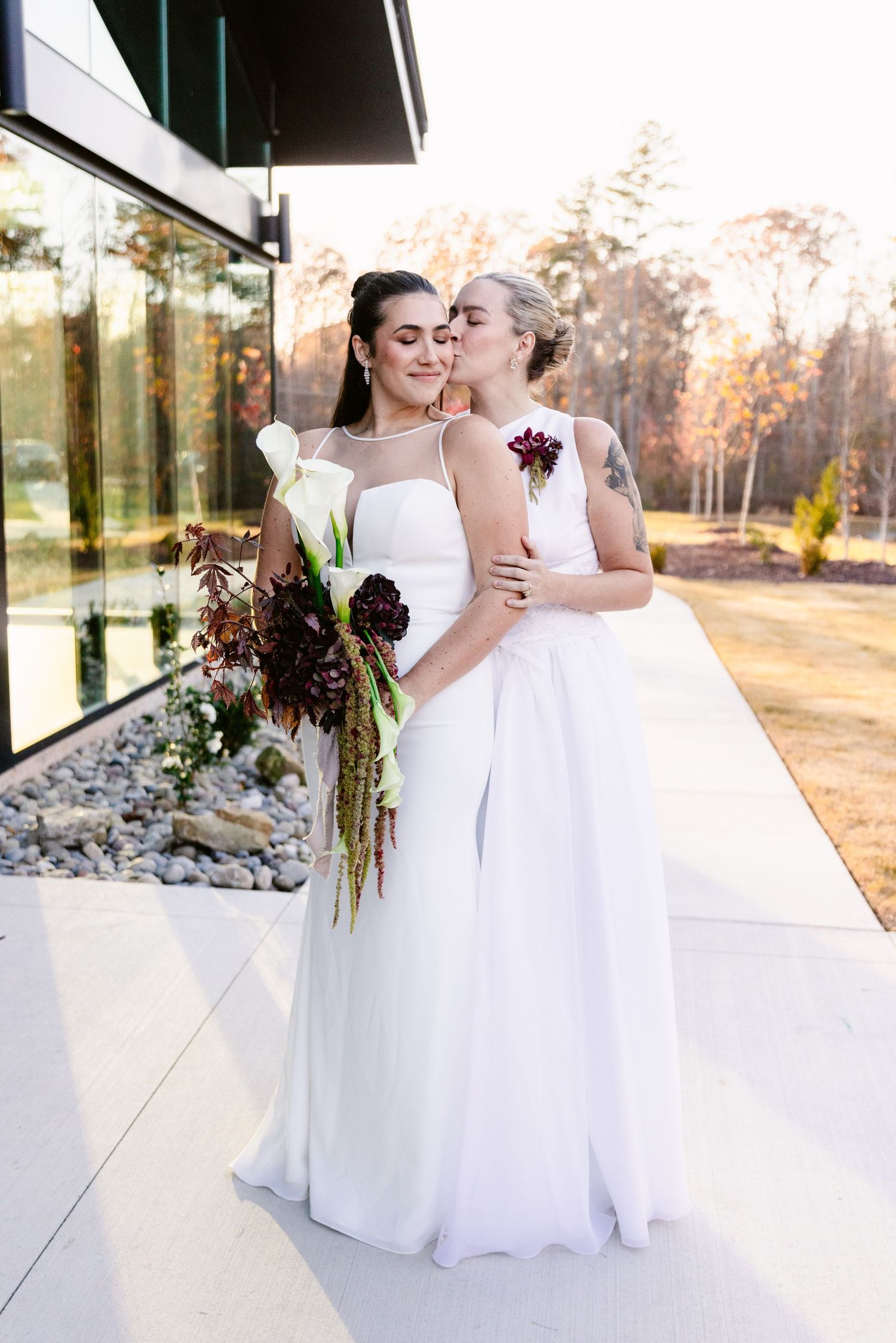 Two people in elegant white wedding dresses share an intimate moment during an outdoor autumn photoshoot.