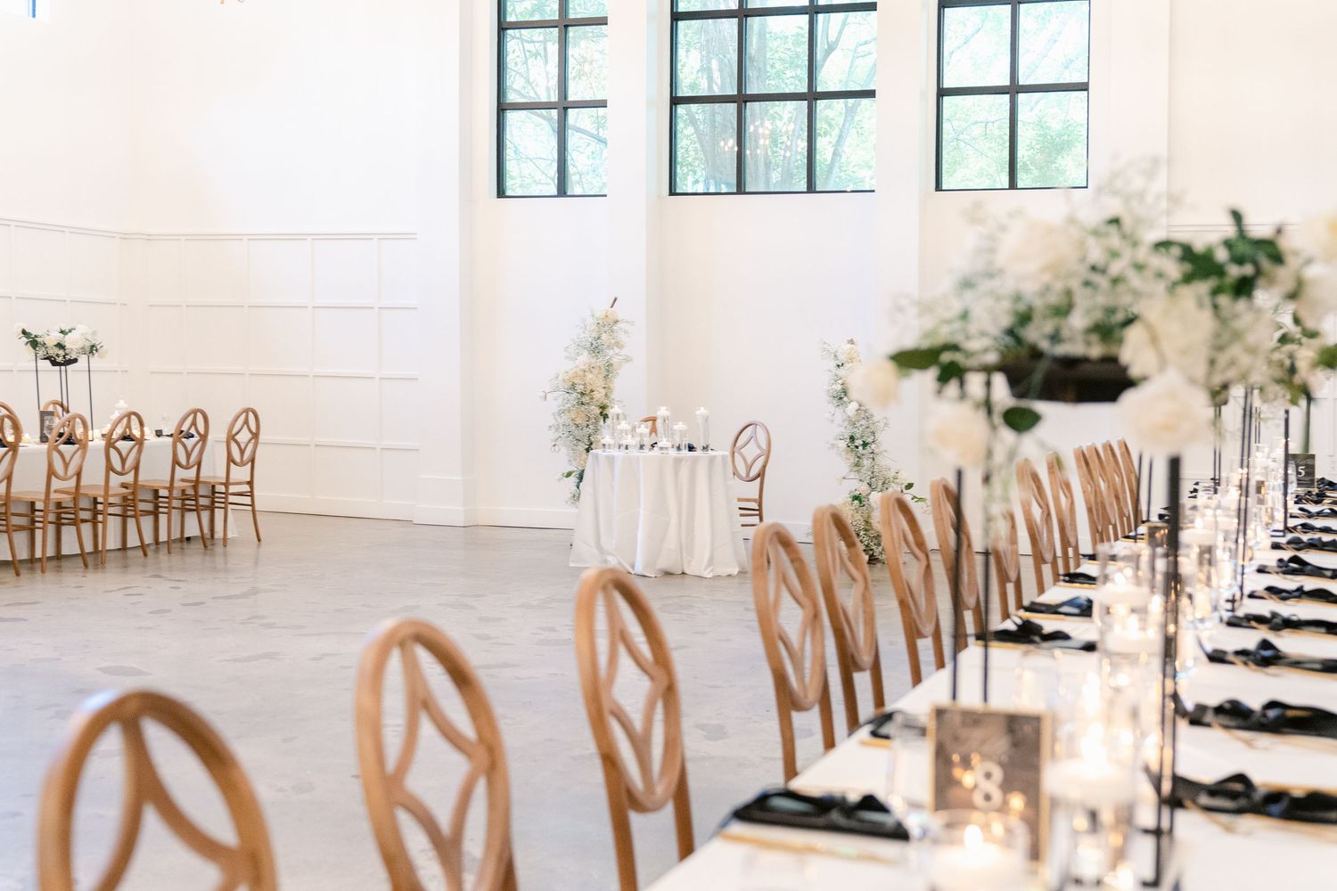 Long banquet tables with white linens and wooden chairs set up in an elegant white room with natural lighting.
