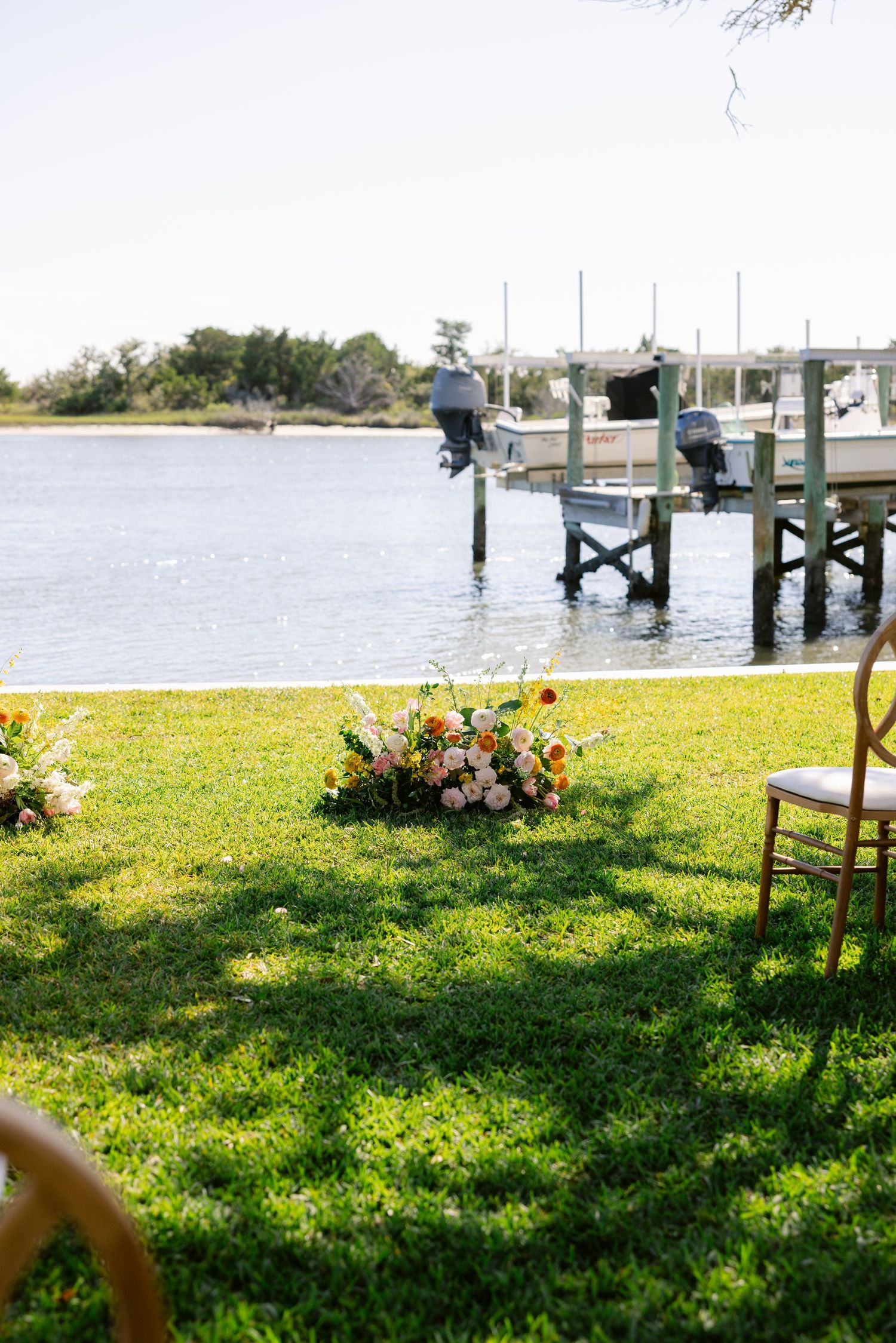 Waterfront wedding setup with floral arrangements on grass near a wooden dock.
