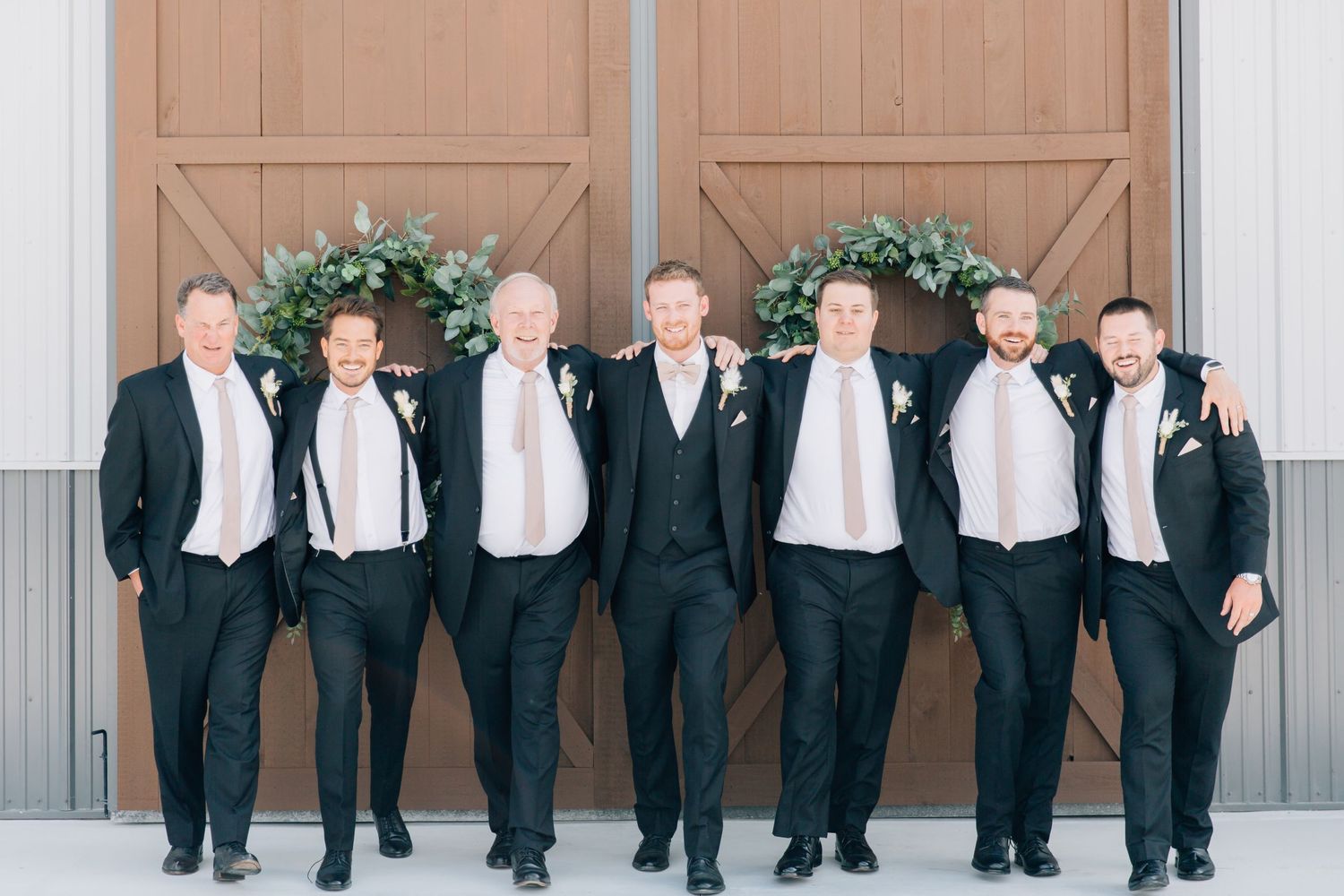 Groomsmen in matching black suits and ties pose together against rustic barn doors.