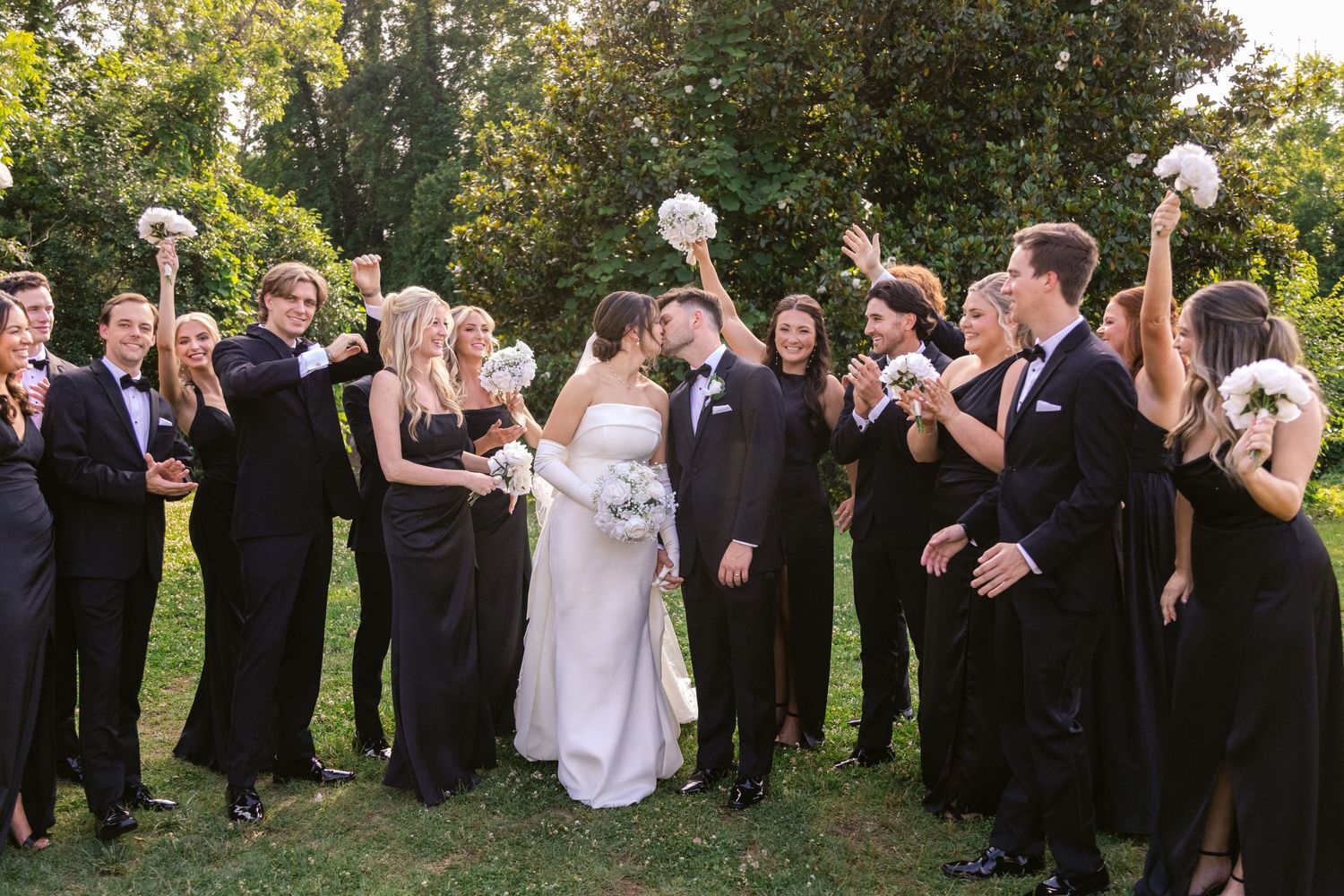 A wedding party in formal black and white attire celebrates outdoors at a sunny garden ceremony.