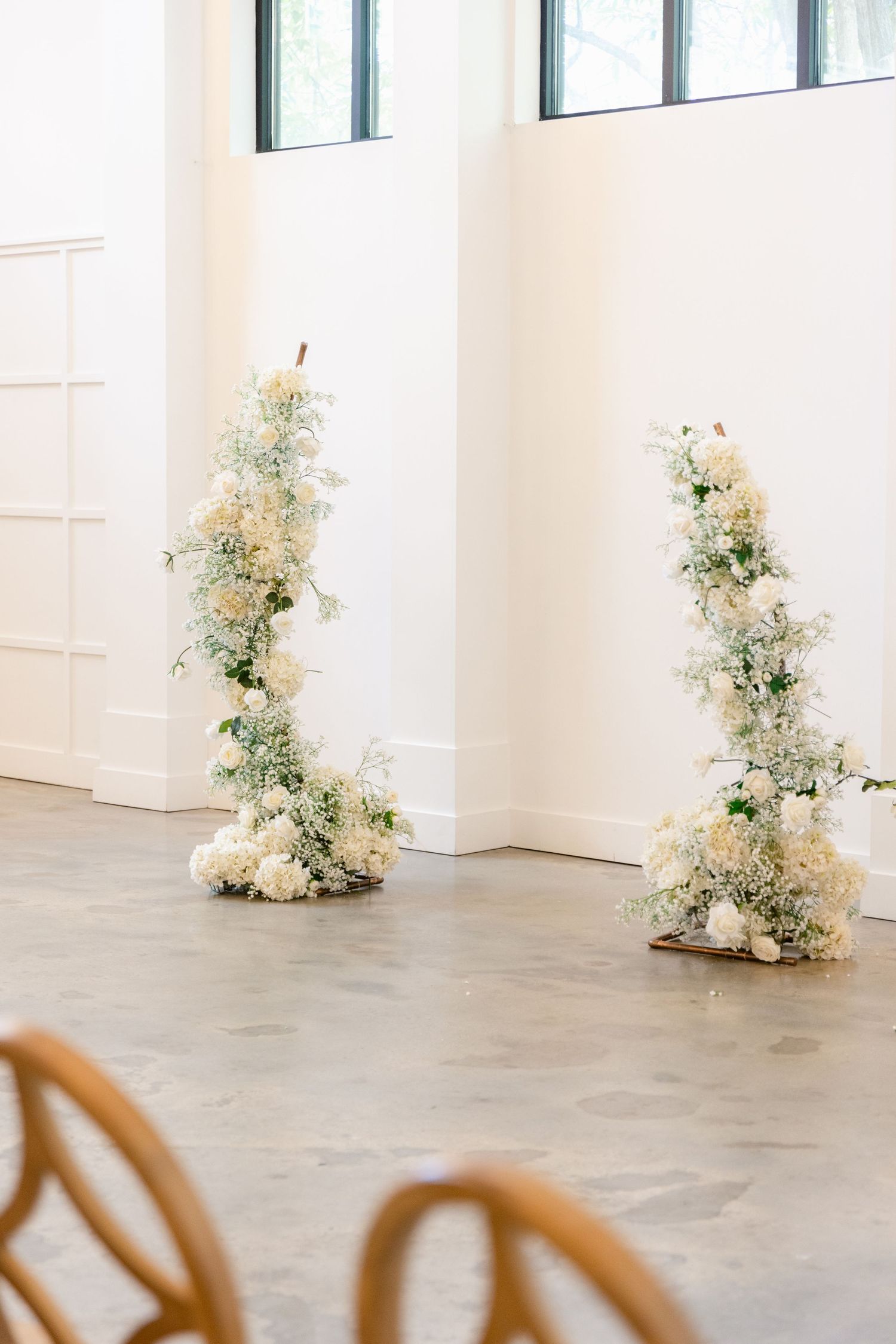 Two elegant white floral arrangements stand tall against a white wall with windows and wooden chairs visible.