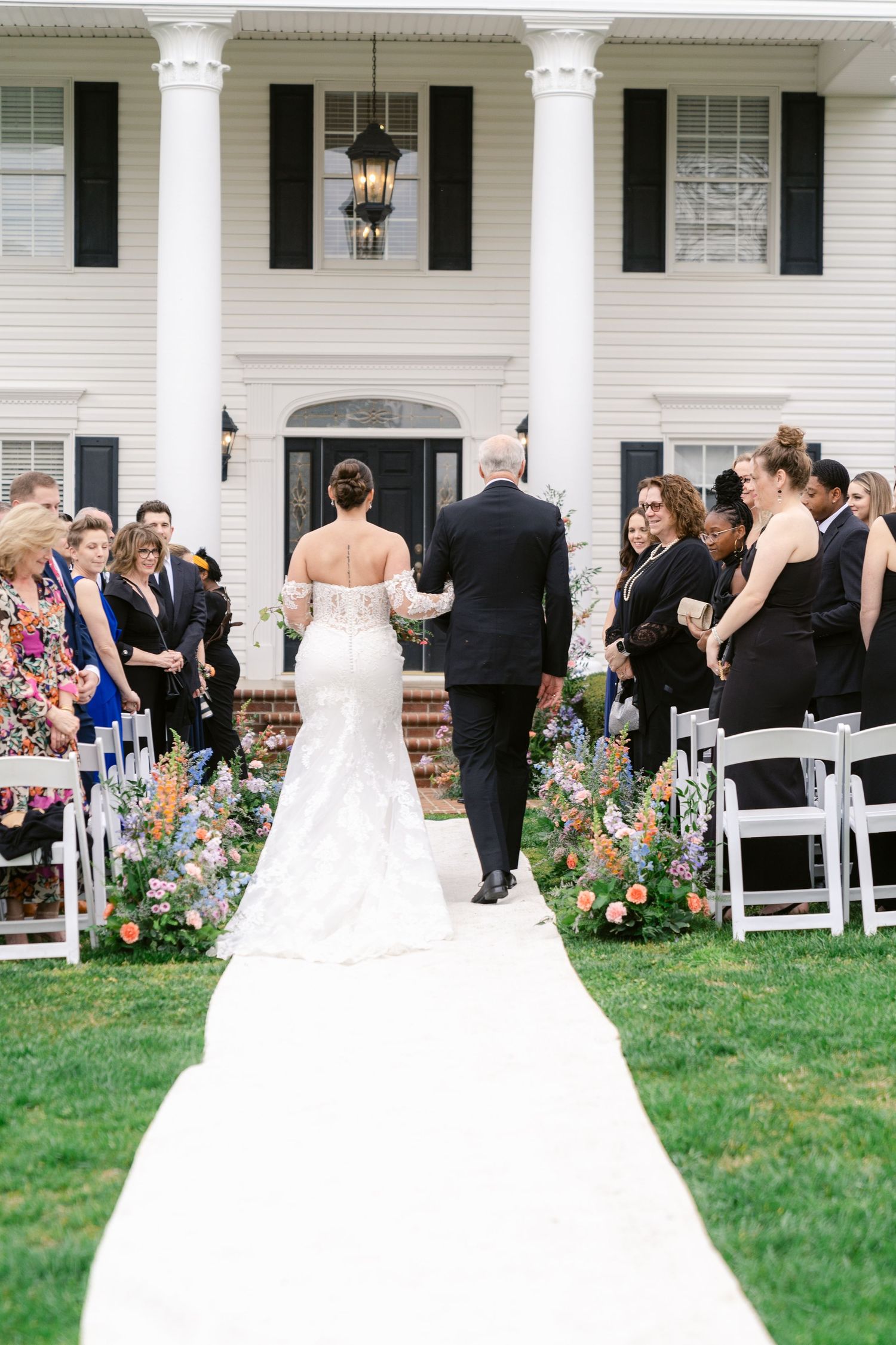 Wedding ceremony setup in front of a grand white colonial house with pillars and floral-lined aisle.