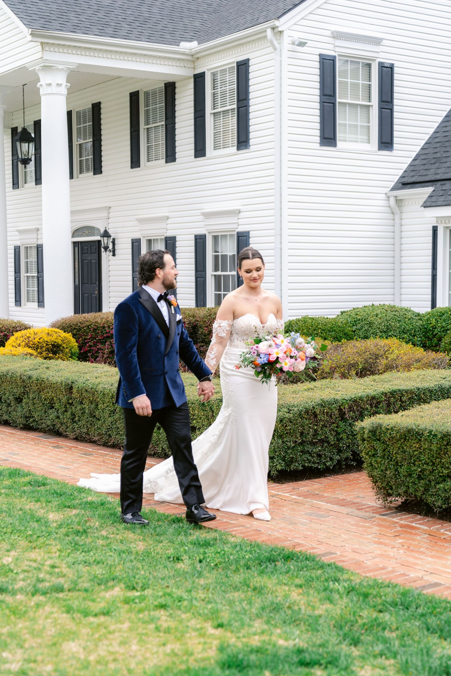A wedding couple stands in front of a classic white colonial house with black shutters and manicured hedges.
