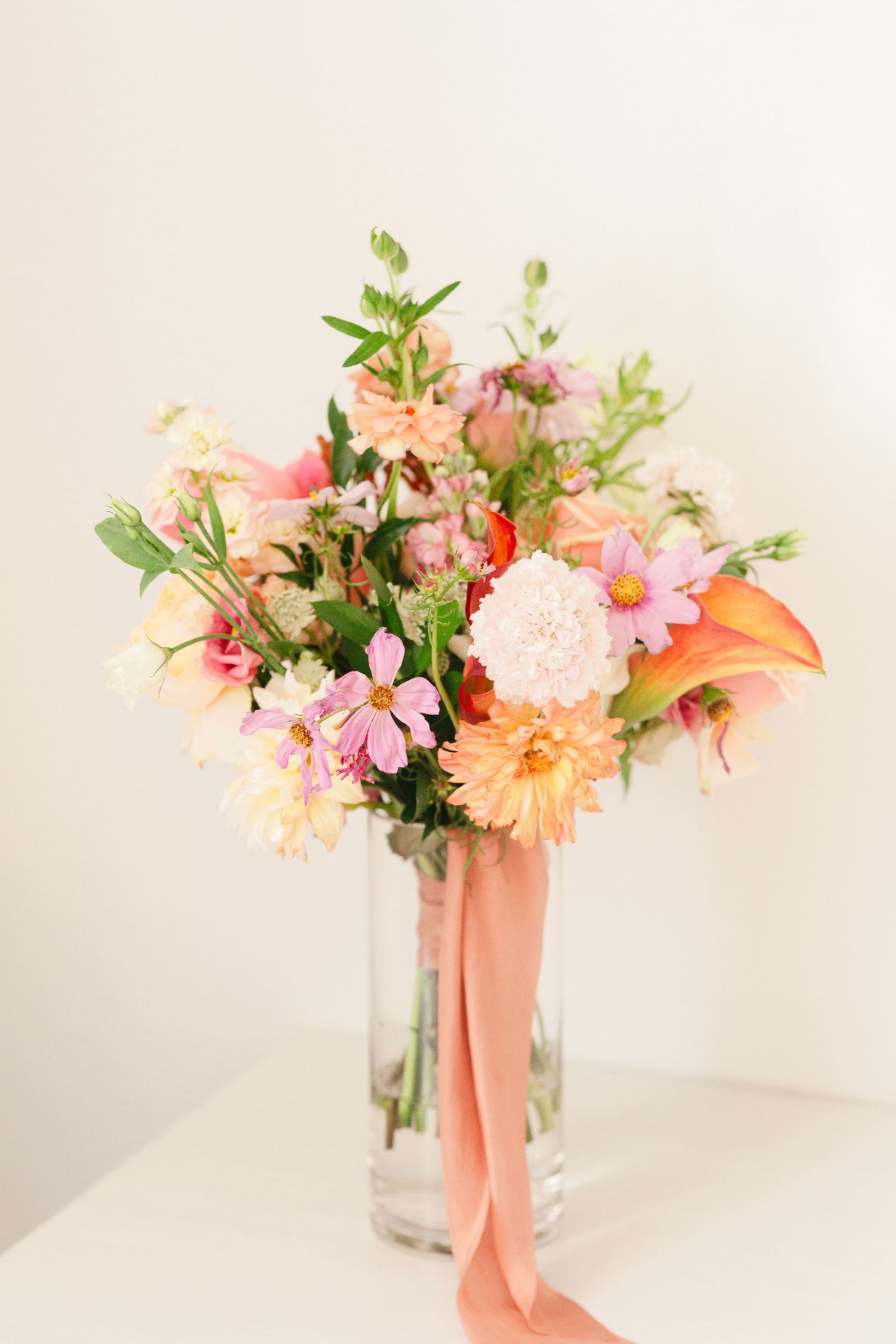 Romantic peach and pink floral arrangement with trailing ribbon in clear glass vase.