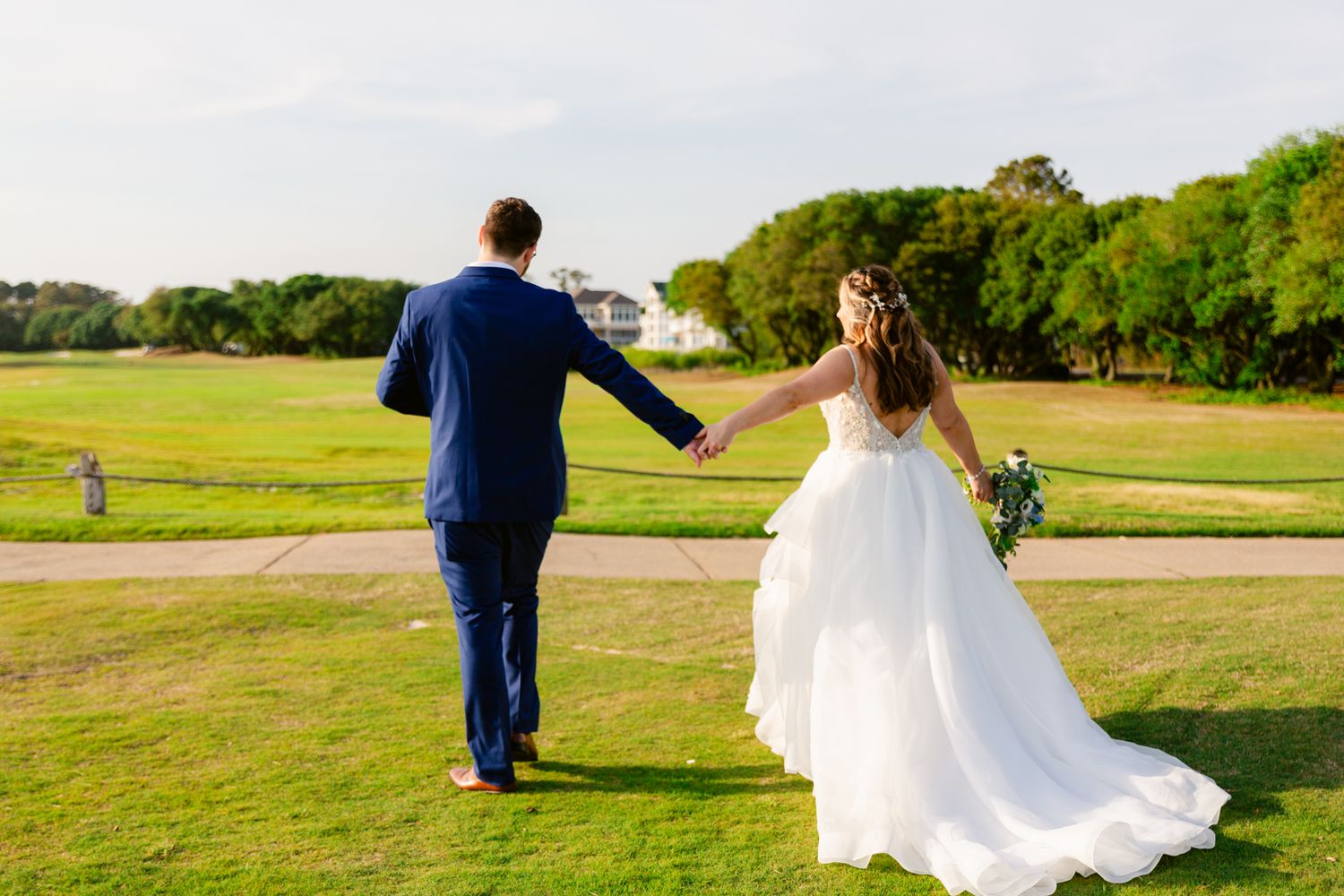 A couple in wedding attire walks hand in hand across a green field on their special day.
