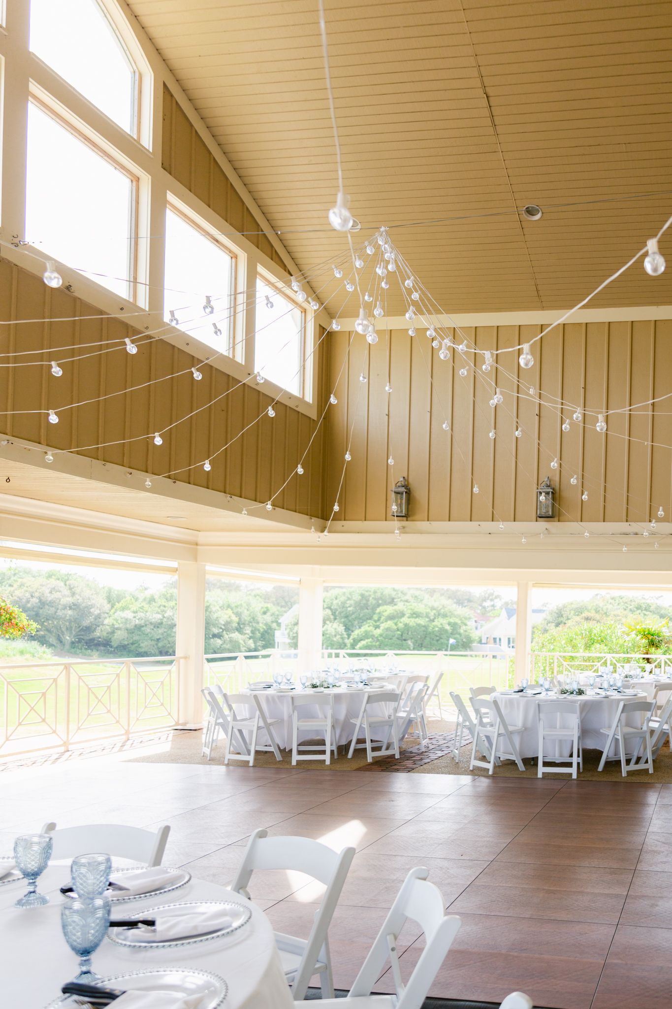 String lights hang from the ceiling of a spacious reception venue with white folding chairs and hardwood floors.