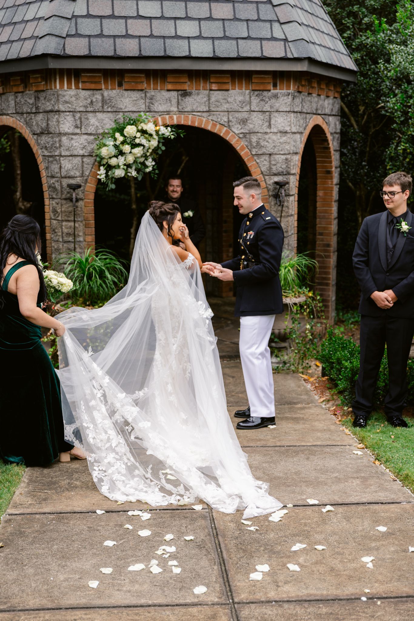A flowing white wedding veil with lace details trails gracefully across a stone courtyard during an outdoor ceremony.