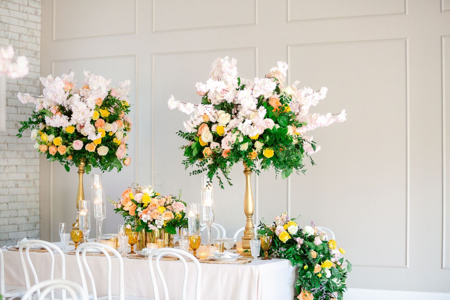 Elegant wedding reception decor features tall floral centerpieces with yellow and white blooms on white tablescapes.