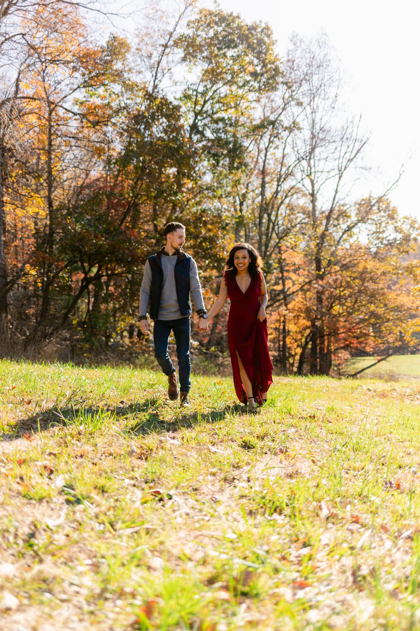 A couple walks hand in hand through a scenic autumn landscape with colorful fall foliage in the background.