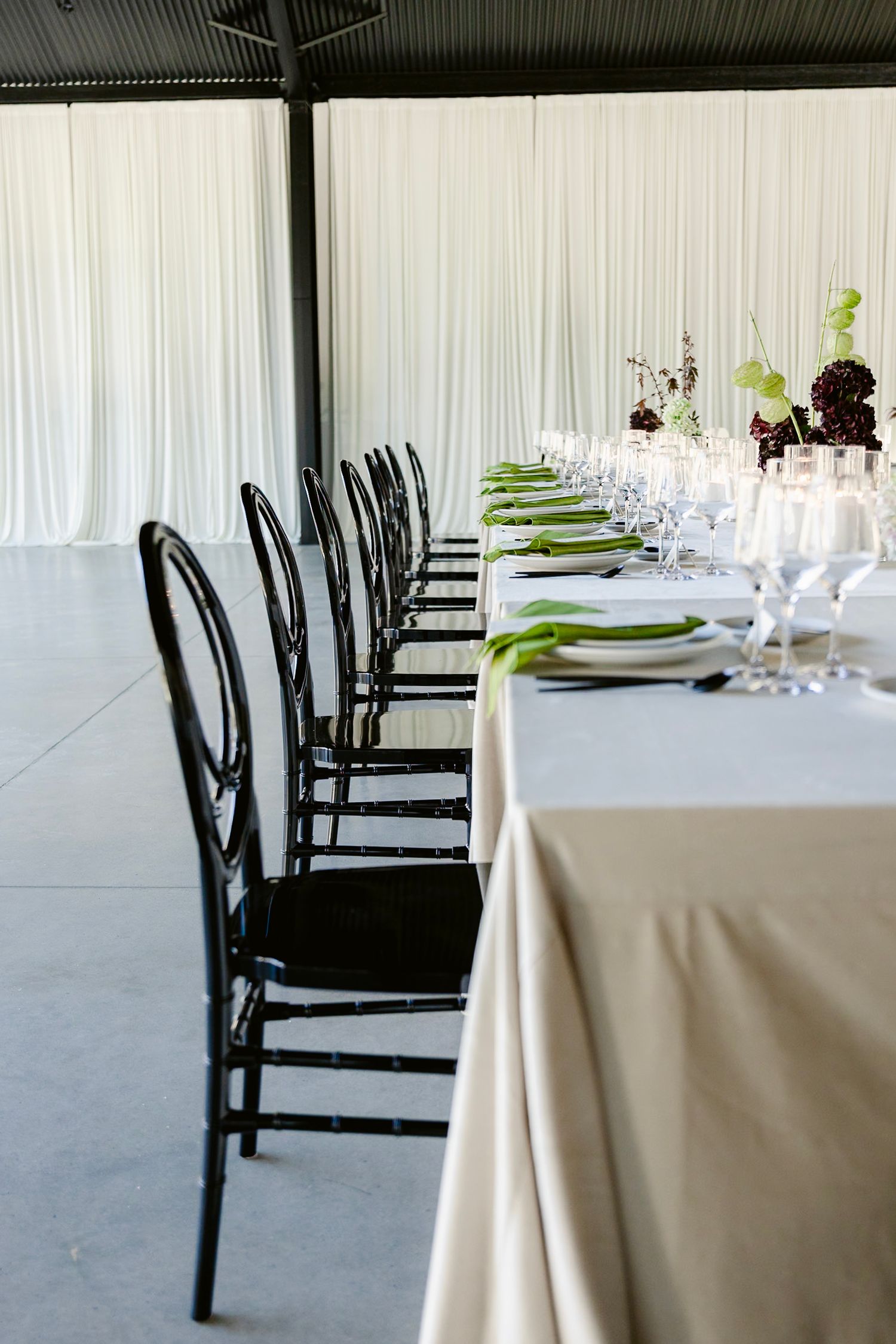 Elegant dining table setup with white tablecloths, black chairs, and green accents in a minimalist event space.