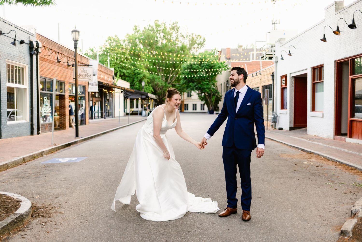 Bride and groom hold hands laughing on a charming brick street lined with shops and string lights on their wedding day.