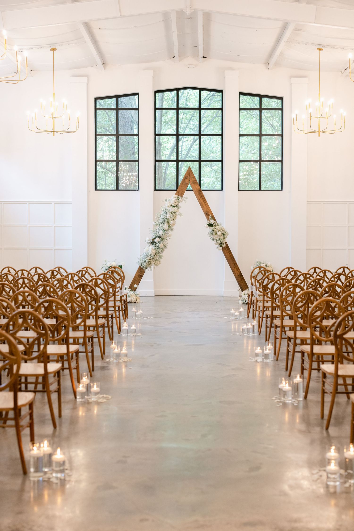 Wooden chairs arranged in rows for a wedding ceremony with candles lining the aisle in a bright white venue.