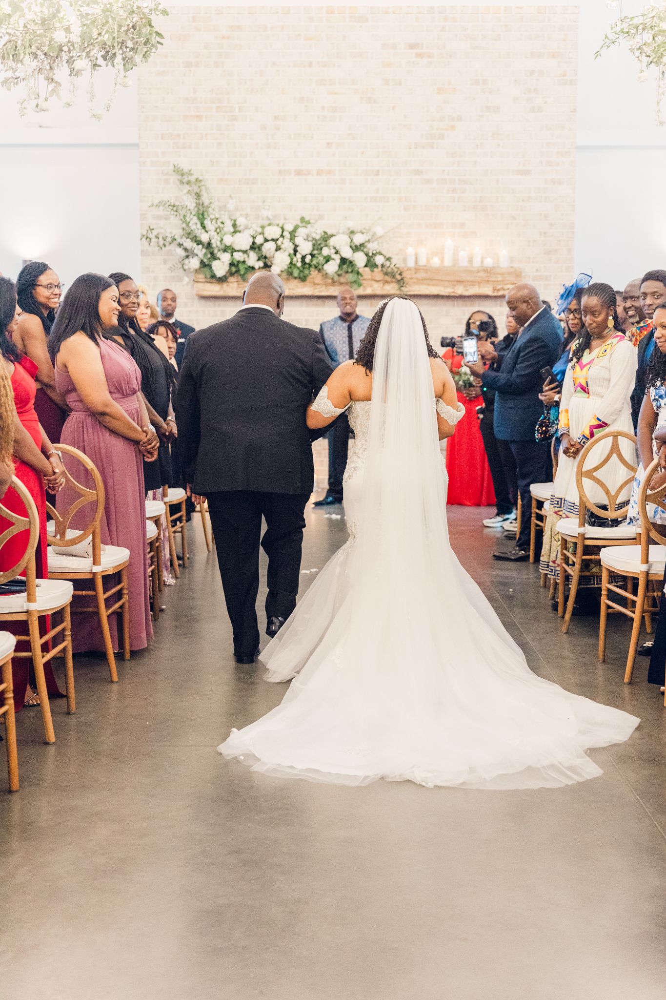 A flowing white wedding dress trails behind as guests look on during an indoor wedding ceremony.