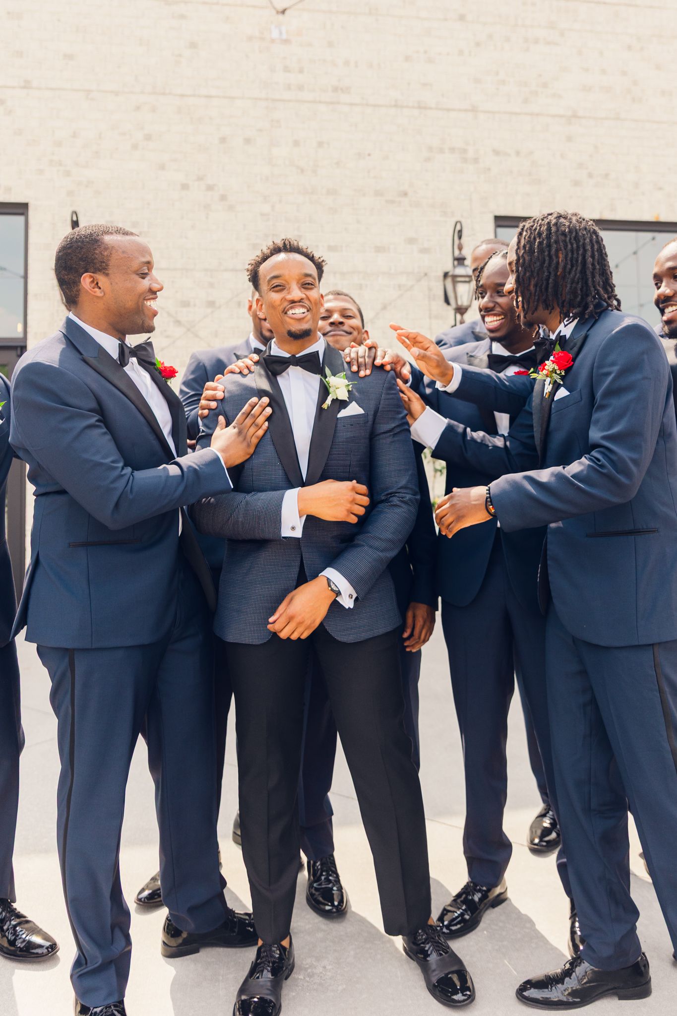 Groomsmen in matching navy suits playfully pose in a line against a white wall during a wedding celebration.