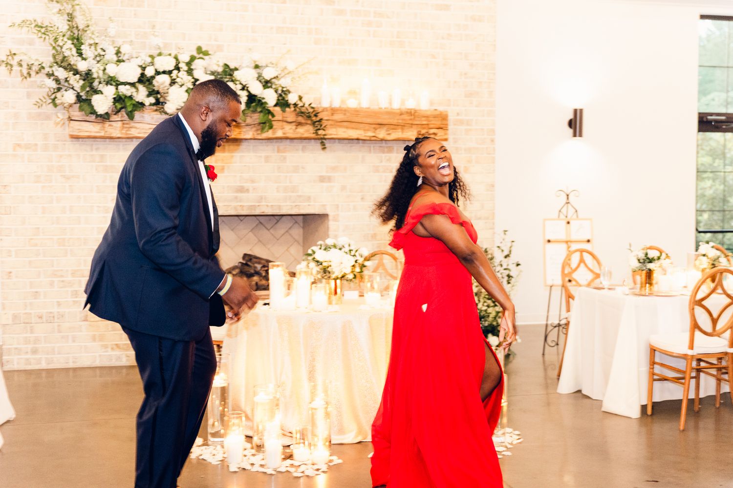 A couple in formal attire dances together in an elegant white reception venue decorated with floral arrangements.