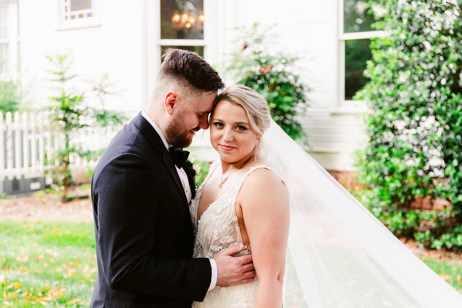 Romantic wedding couple embraces in garden setting with white house and veil in background.
