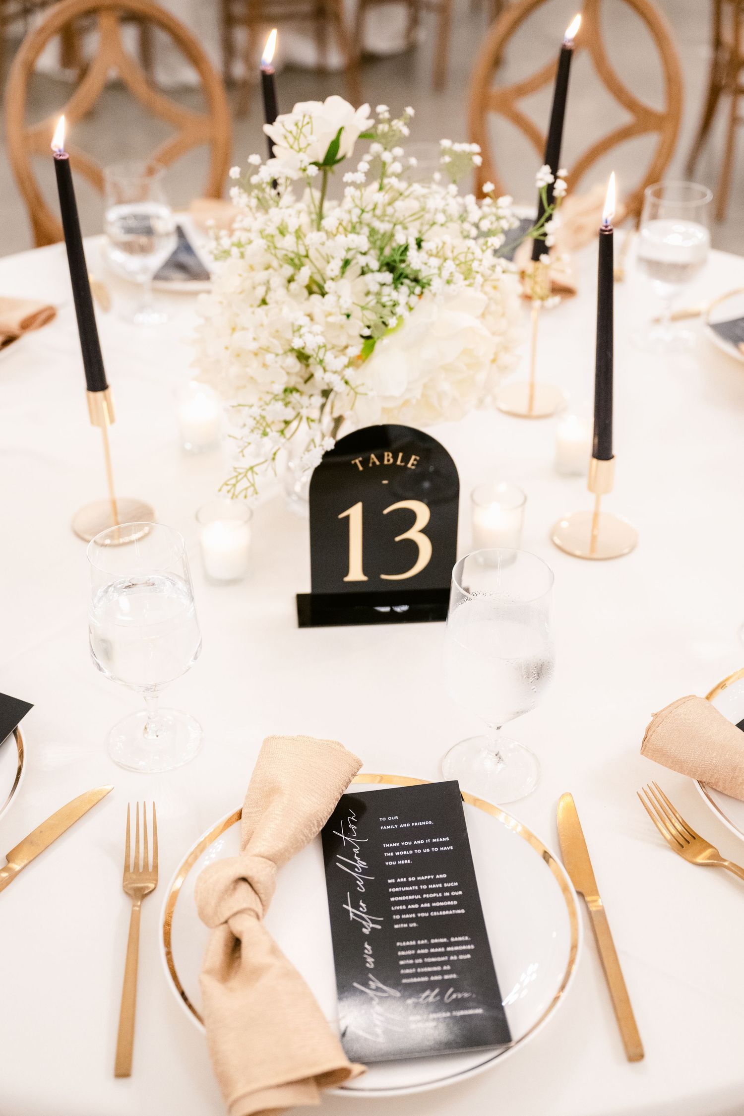 Elegant wedding table setting with black menu cards, gold cutlery, and white floral centerpieces against a crisp white tablecloth.