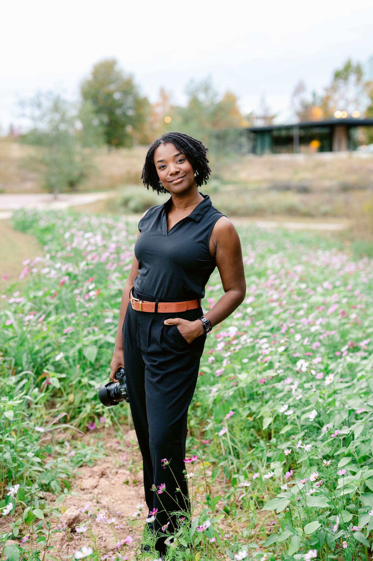 A photographer in a black outfit poses with their camera in a field of wildflowers during golden hour.