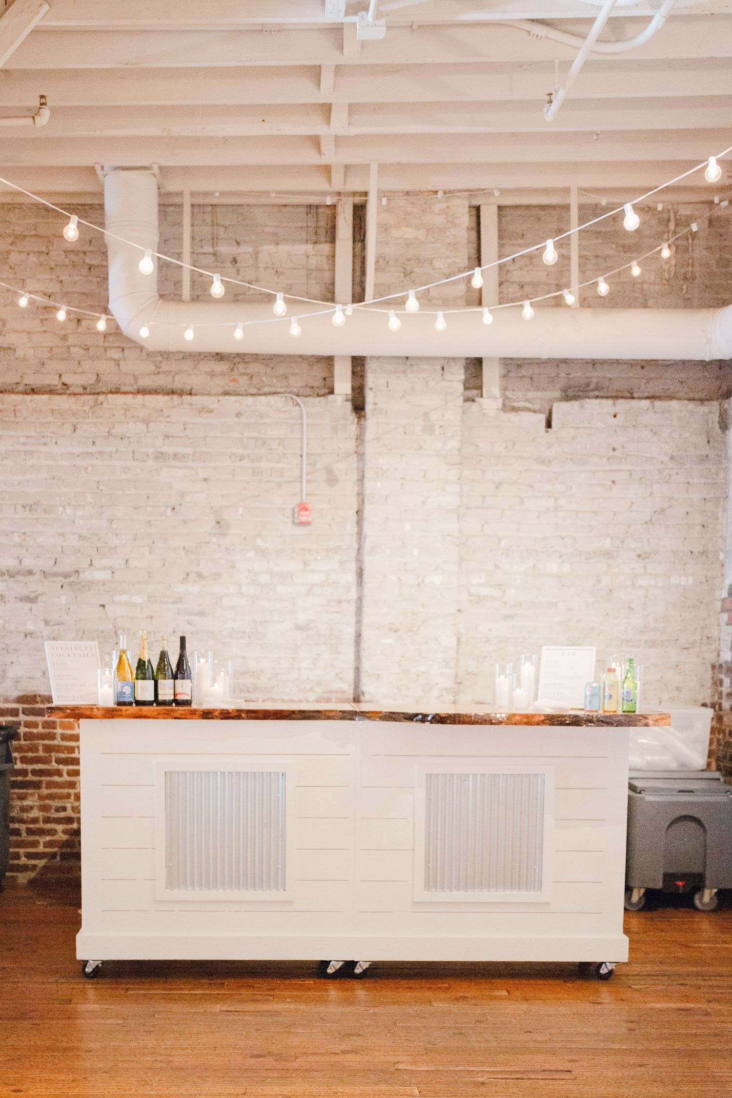 White mobile bar cart with string lights and bottles displayed in industrial wedding venue.