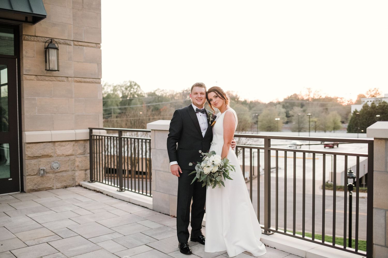 Formal wedding portrait on stone balcony overlooking scenic water view at dusk.