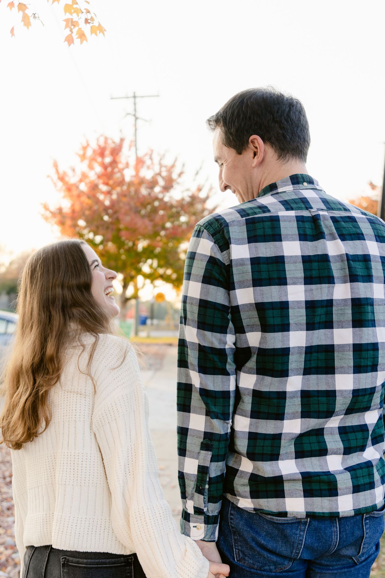 A couple in autumn, wearing a plaid flannel shirt and cream sweater, stand together against fall foliage.